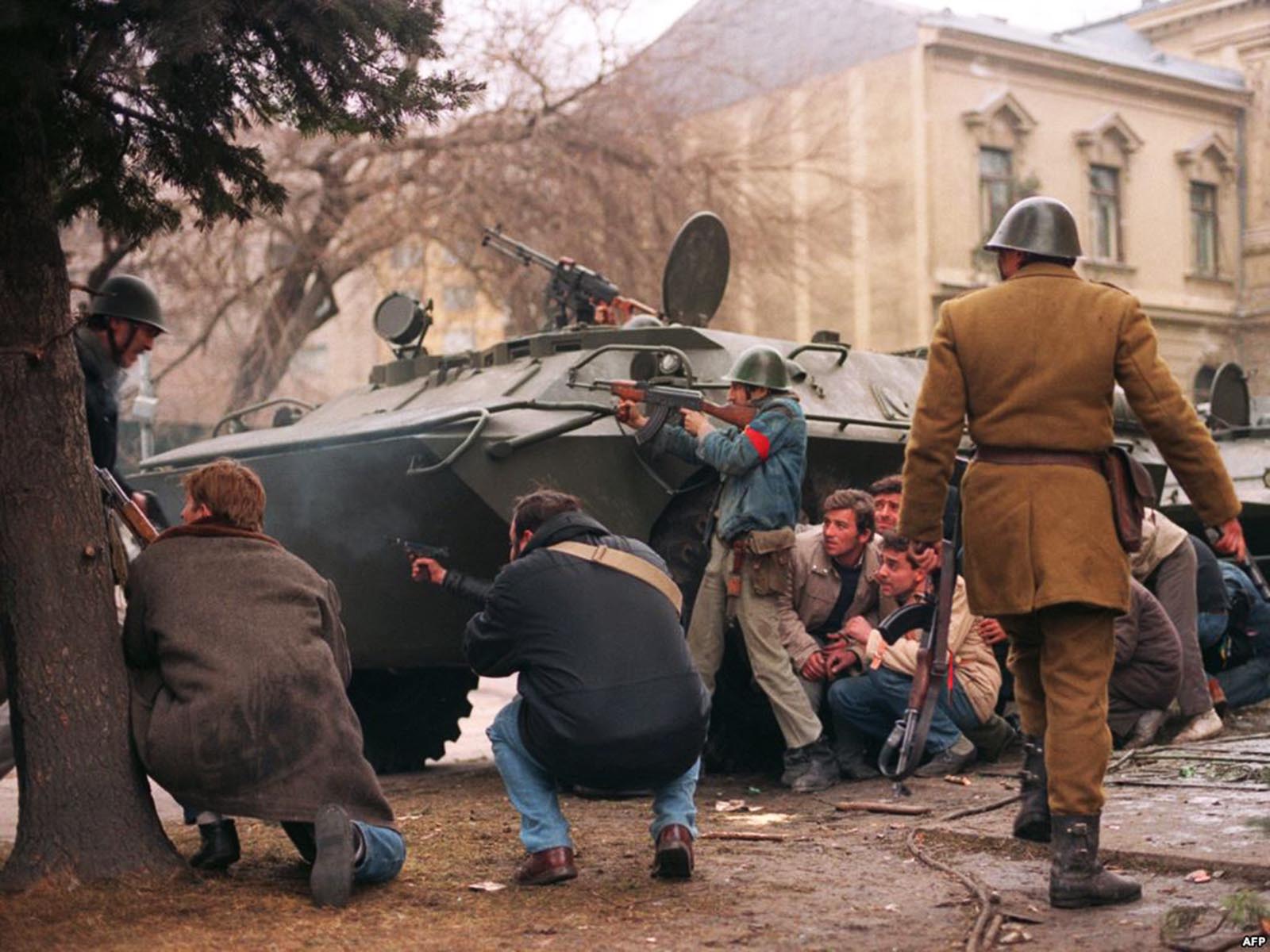 A group of anti-Communist civilian fighters along with a Romanian soldier supporting anti-Ceausescu's activists, protect themselves behind an armoured personnel carrier from snipers fire shot by the supposedly Securitate secret police agents loyal to the communist power .