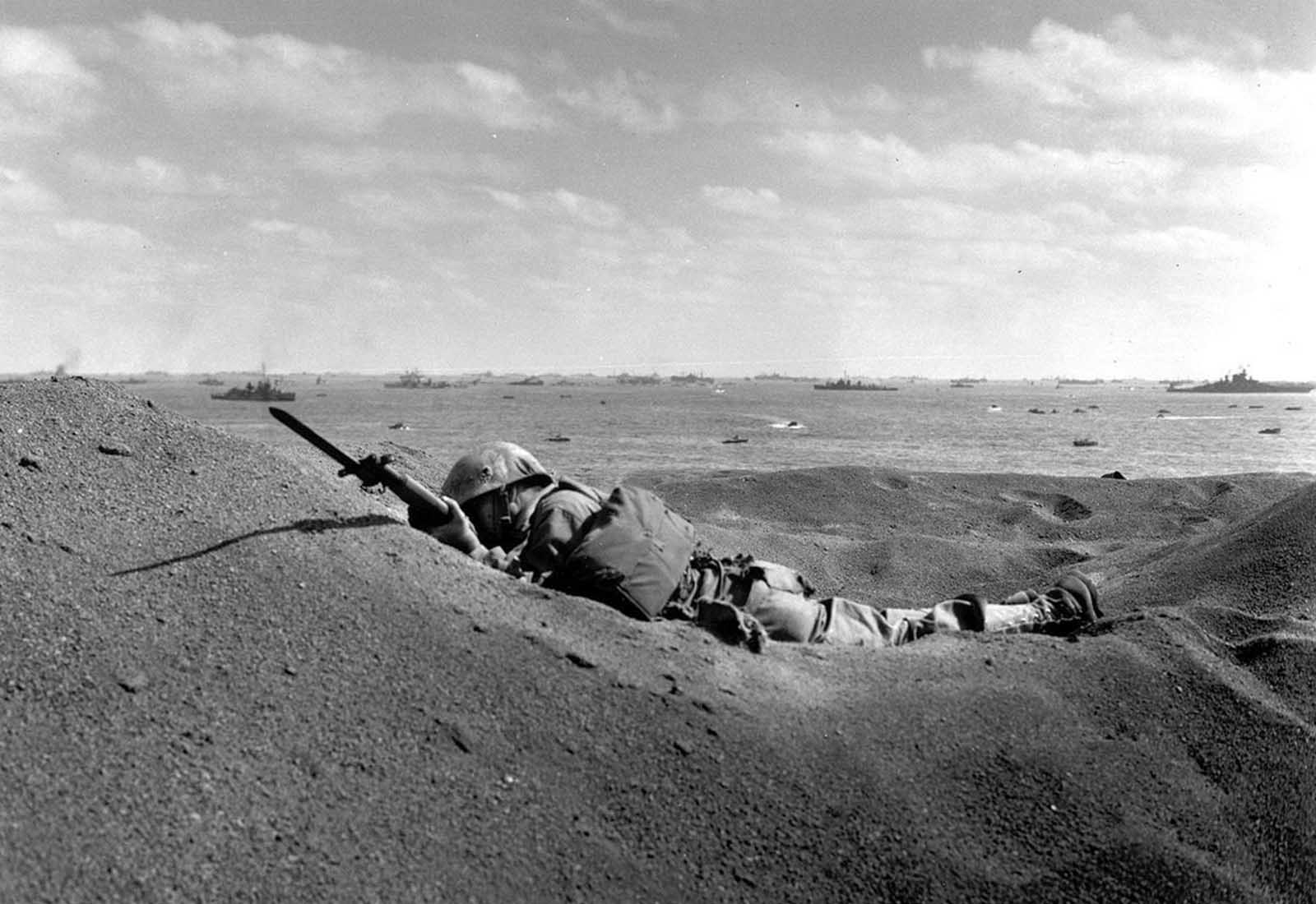 A U.S. Marine, killed by Japanese sniper fire, still holds his weapon as he lies in the black volcanic sand of Iwo Jima, on February 19, 1945, during the initial invasion on the island. In the background are the battleships of the U.S. fleet that made up the invasion task force.