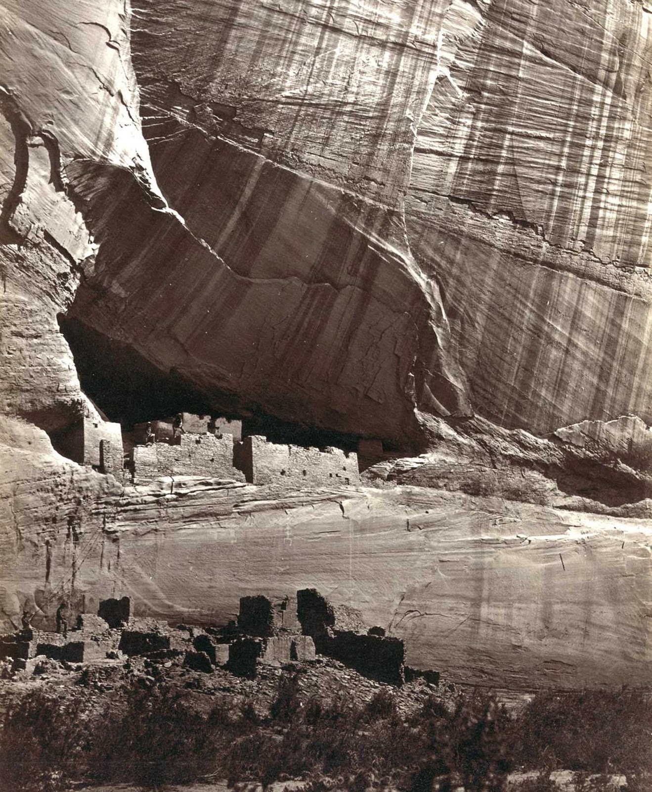 View of the White House, Ancestral Pueblo Native American (Anasazi) ruins in Canyon de Chelly, Arizona, in 1873. The cliff dwellings were built by the Anasazi more than 500 years earlier. At bottom, men stand and pose on cliff dwellings in a niche and on ruins on the canyon floor. Climbing ropes connect the groups of men.