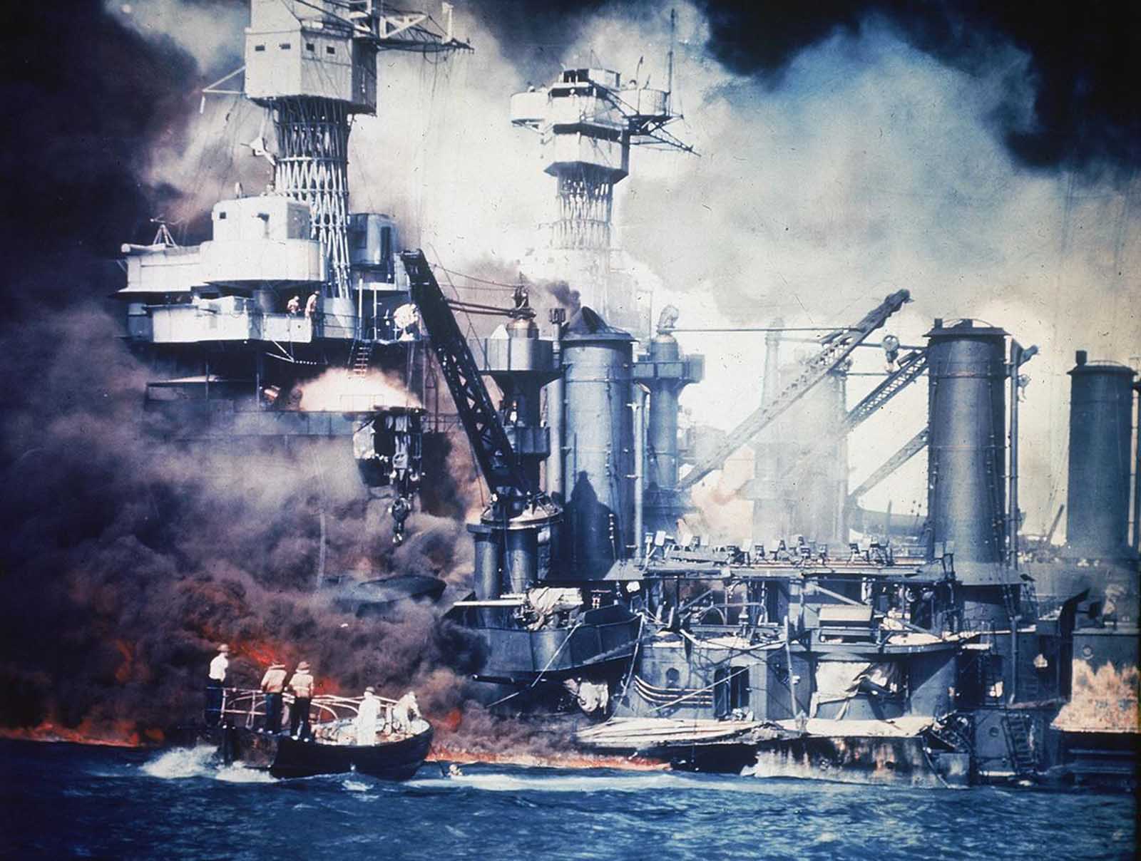 A small boat rescues a USS West Virginia crew member from the water after the Japanese bombing of Pearl Harbor, Hawaii, on December 7, 1941. Two men can be seen on the superstructure, upper center. The mast of the USS Tennessee is beyond the burning West Virginia.