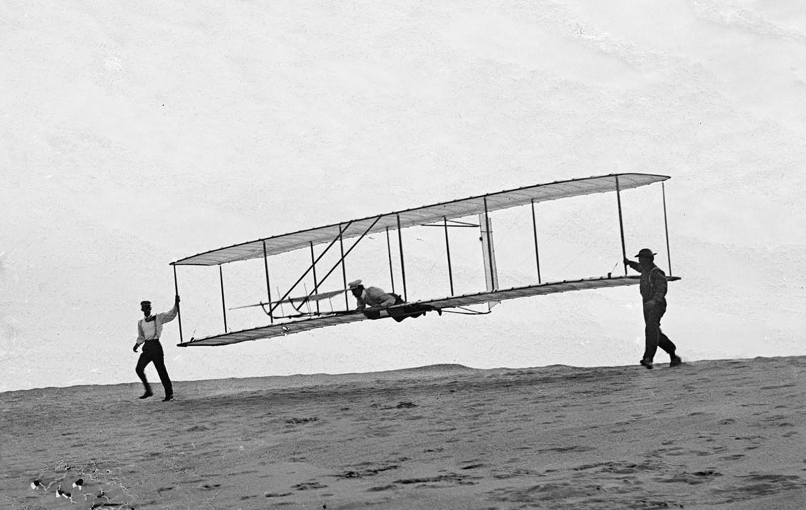 Start of a glide; Wilbur in motion at left holding one end of glider (rebuilt with single vertical rudder), Orville lying prone in machine, and Dan Tate at right, in Kitty Hawk, North Carolina, on October 10, 1902.