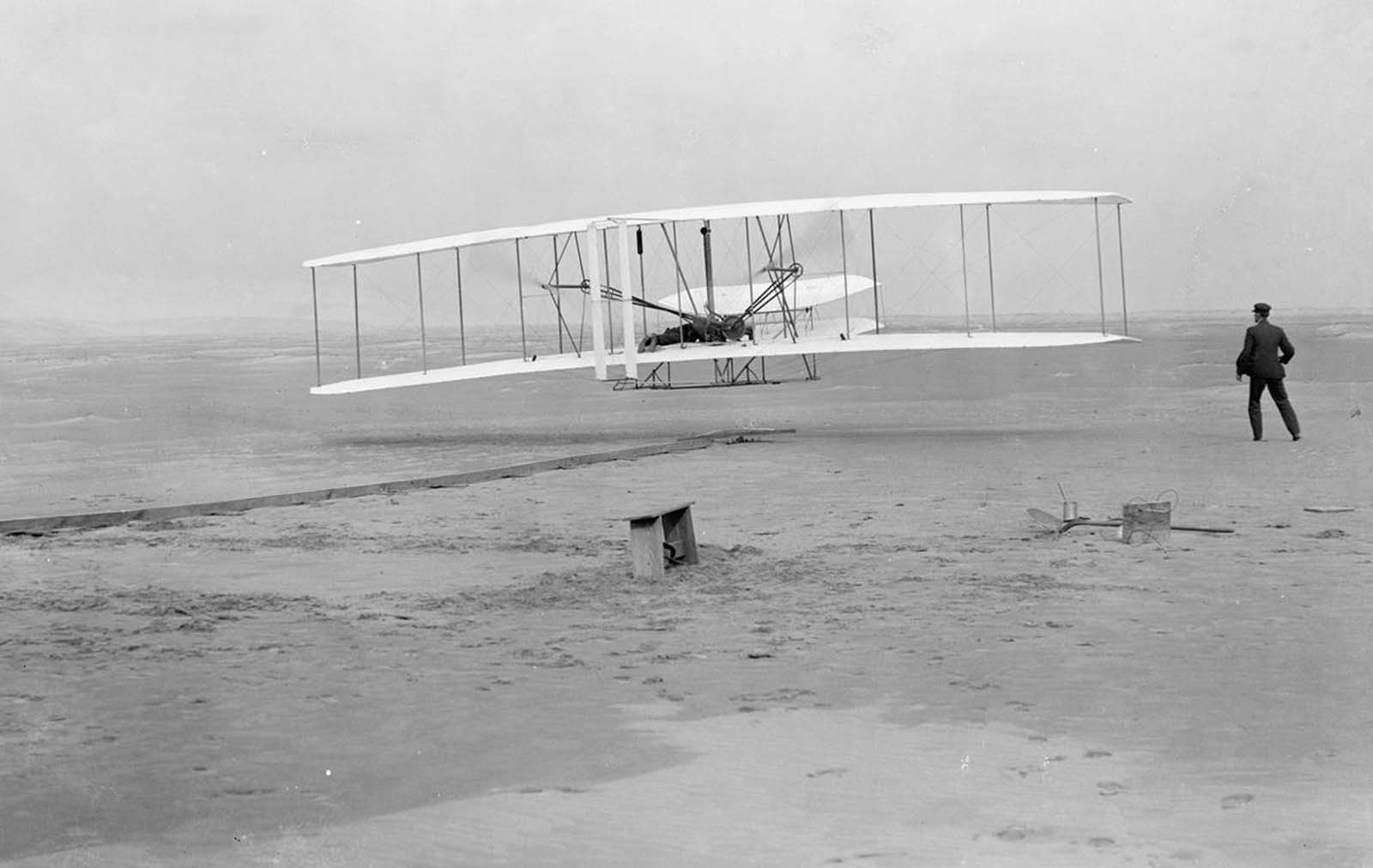 First flight: 120 feet in 12 seconds, on December 17, 1903. This photograph shows man's first powered, controlled, sustained flight. Orville Wright at the controls of the machine, lying prone on the lower wing with hips in the cradle which operated the wing-warping mechanism. Wilbur Wright running alongside to balance the machine, has just released his hold on the forward upright of the right wing. The starting rail, the wing-rest, a coil box, and other items needed for flight preparation are visible behind the machine. Orville Wright preset the camera and had John T. Daniels squeeze the rubber bulb, tripping the shutter.
