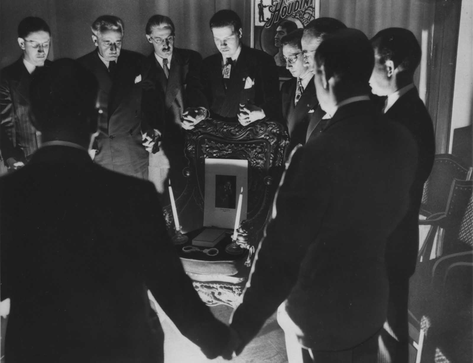 Thirteen magicians gather around a chair on which are a portrait of Houdini, two candles, a book, and handcuffs, for a midnight seance on Halloween, 1946, in Detroit, Michigan.