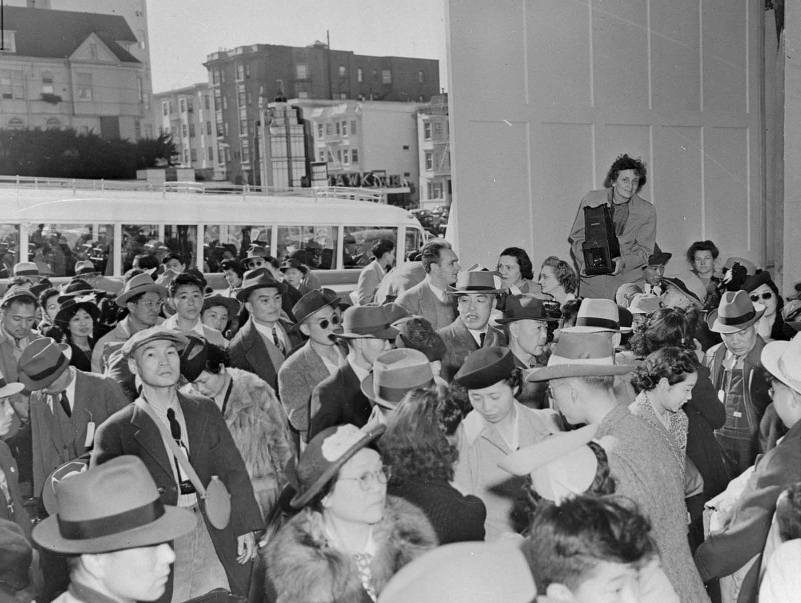 Japanese-Americans assemble in San Francisco for transportation to an assembly center, and later to various relocation centers. Photographer Dorothea Lange is visible above the crowd.