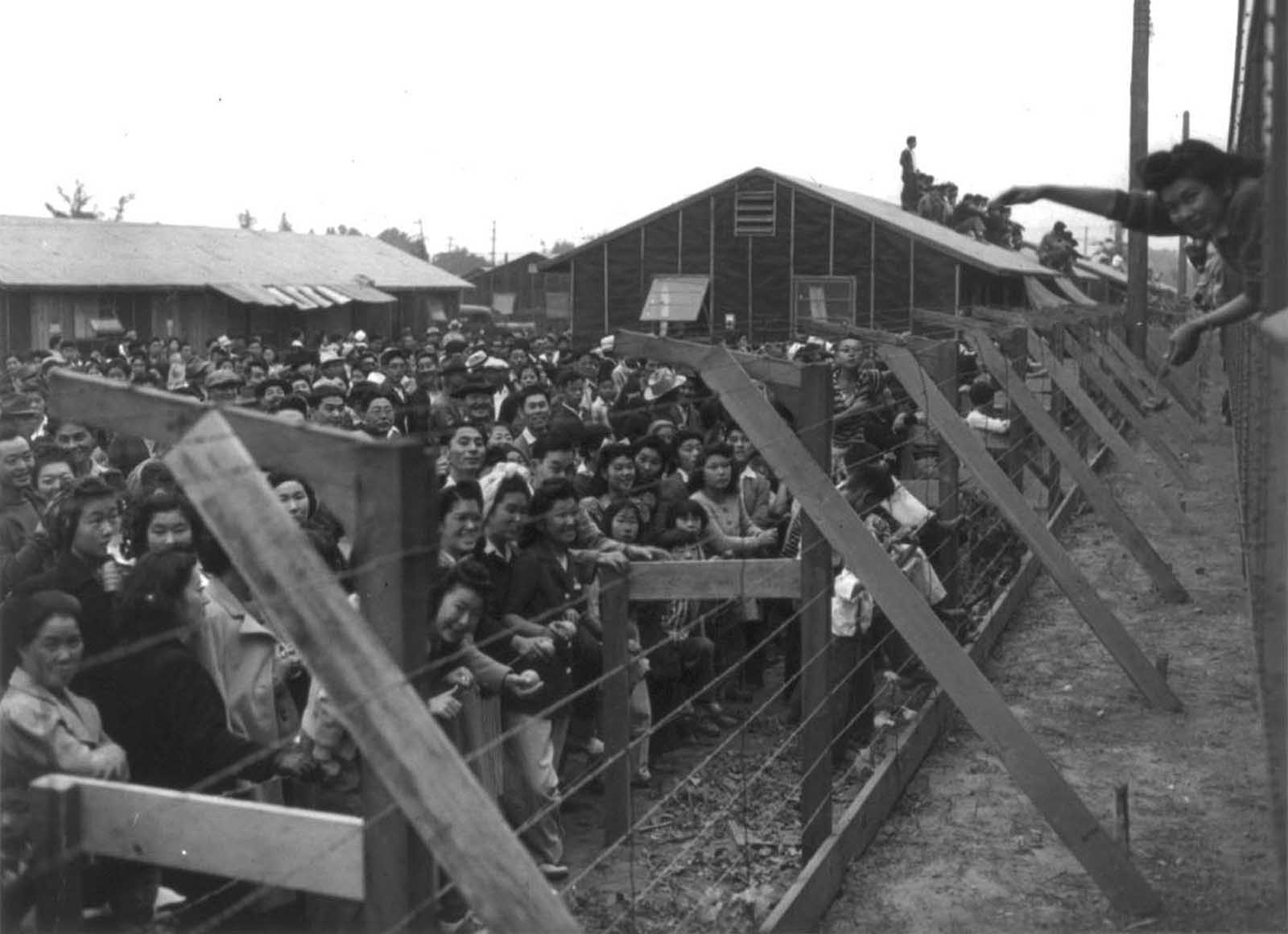 A crowd of Japanese Americans stand behind a barbed wire fence waving to departing friends on train leaving Santa Anita, California.