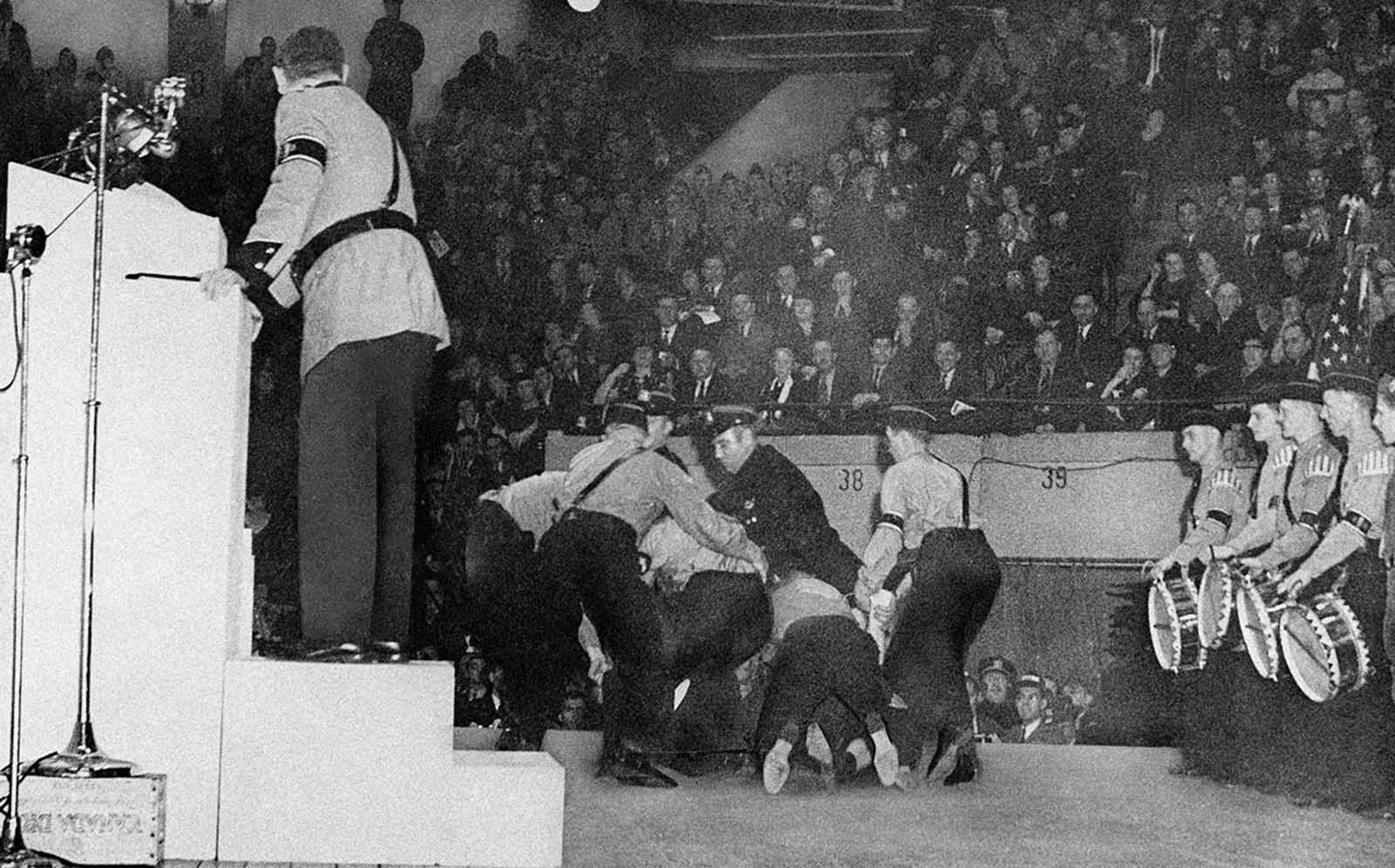 Stormtroopers subdue a heckler on the platform at New York’s Madison Square Garden, February 20, 1939. Police who rescued and later arrested the man, whose clothing was torn from him in the struggle, identified him as Isadore Greenbaum, 26, a hotel worker. Fritz Kuhn, National Bund leader, stands on the rostrum, his back turned as he regards the struggle which interrupted his Denunciation of Jews during the Bund rally.