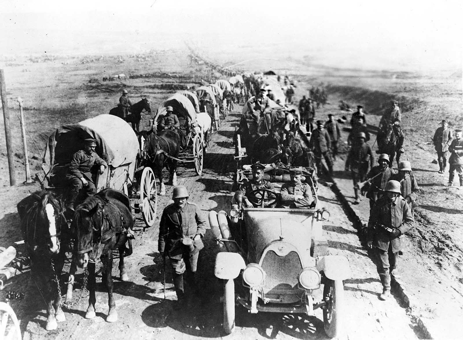 German officers in an automobile on the road with a convoy of wagons; soldiers walk along side the road.