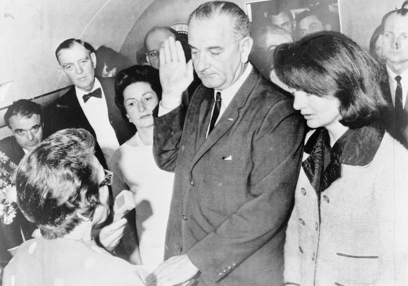 Lyndon B. Johnson is sworn in as President by Federal District Judge Sarah T. Hughes aboard the Presidential plane, as wife Lady Bird, former First Lady Jacqueline Kennedy and others look on.