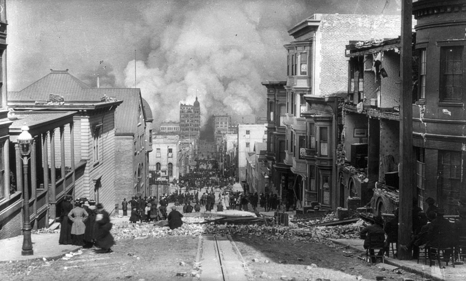 San Francisco residents, some seated in chairs, sit among the earthquake damage, watching out-of-control fires in the distance.