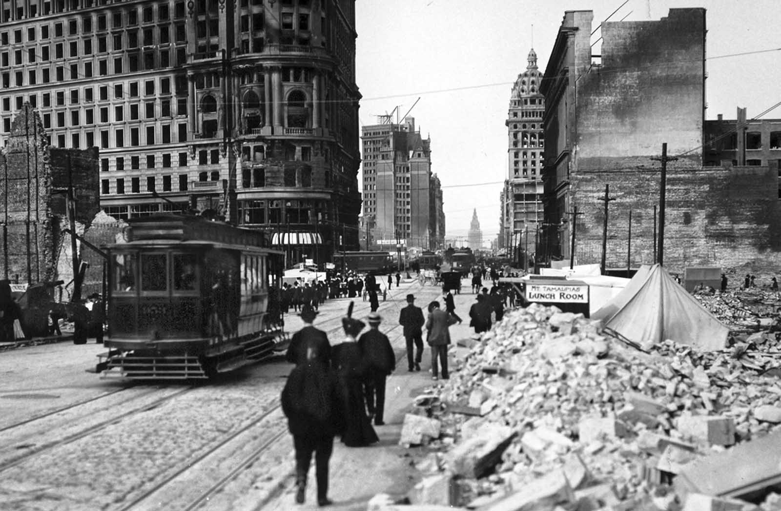Market Street at the junction of Powell and Market, looking east toward the Ferry Building. The large building to the left is the Flood Building. The side wall of the first large building is the Emporium, the largest department store in San Francisco. On the right is the San Francisco Call Building. On the left, the structure with the derrick is the new addition to the De Young Building, and in the extreme distance, at the end of Market Street, is the tower of the Ferry Building.
