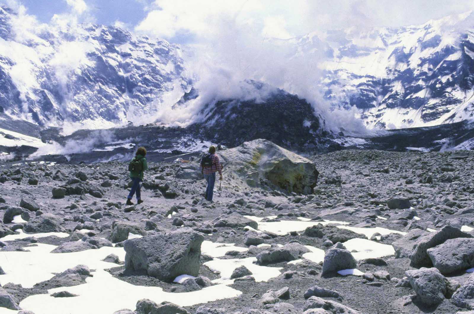 A Washington state geologist and a reporter walk toward the crater of Mount St. Helens in May of 1981, almost a year after the volcano exploded, causing dramatic destruction for miles around. They were near the steaming lava dome with the snow-covered crater wall beyond.