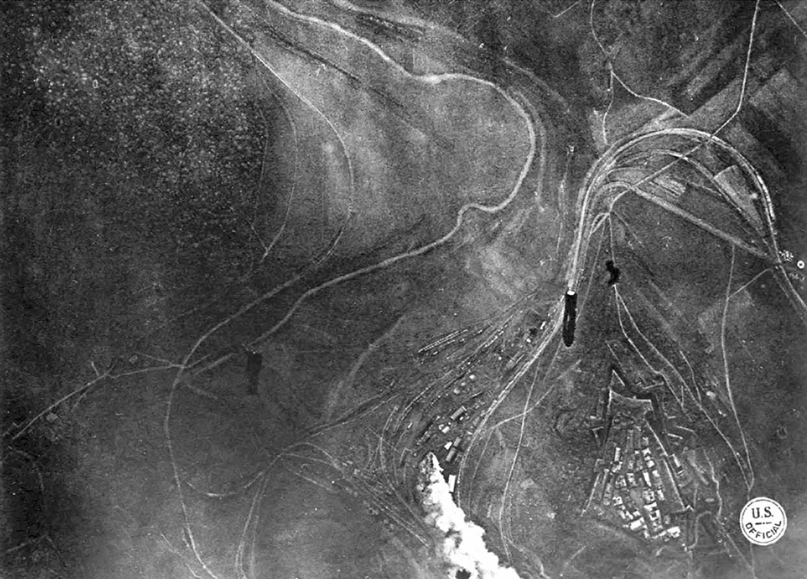 Bombing Montmedy, 42 km north of Verdun, while American troops advance in the Meuse-Argonne sector. Three bombs have been released by a U.S. bomber, one striking a supply station, the other two in mid-air, visible on their way down. Black puffs of smoke indicate anti-aircraft fire. To the right (west), a building with a Red Cross symbol can be seen.