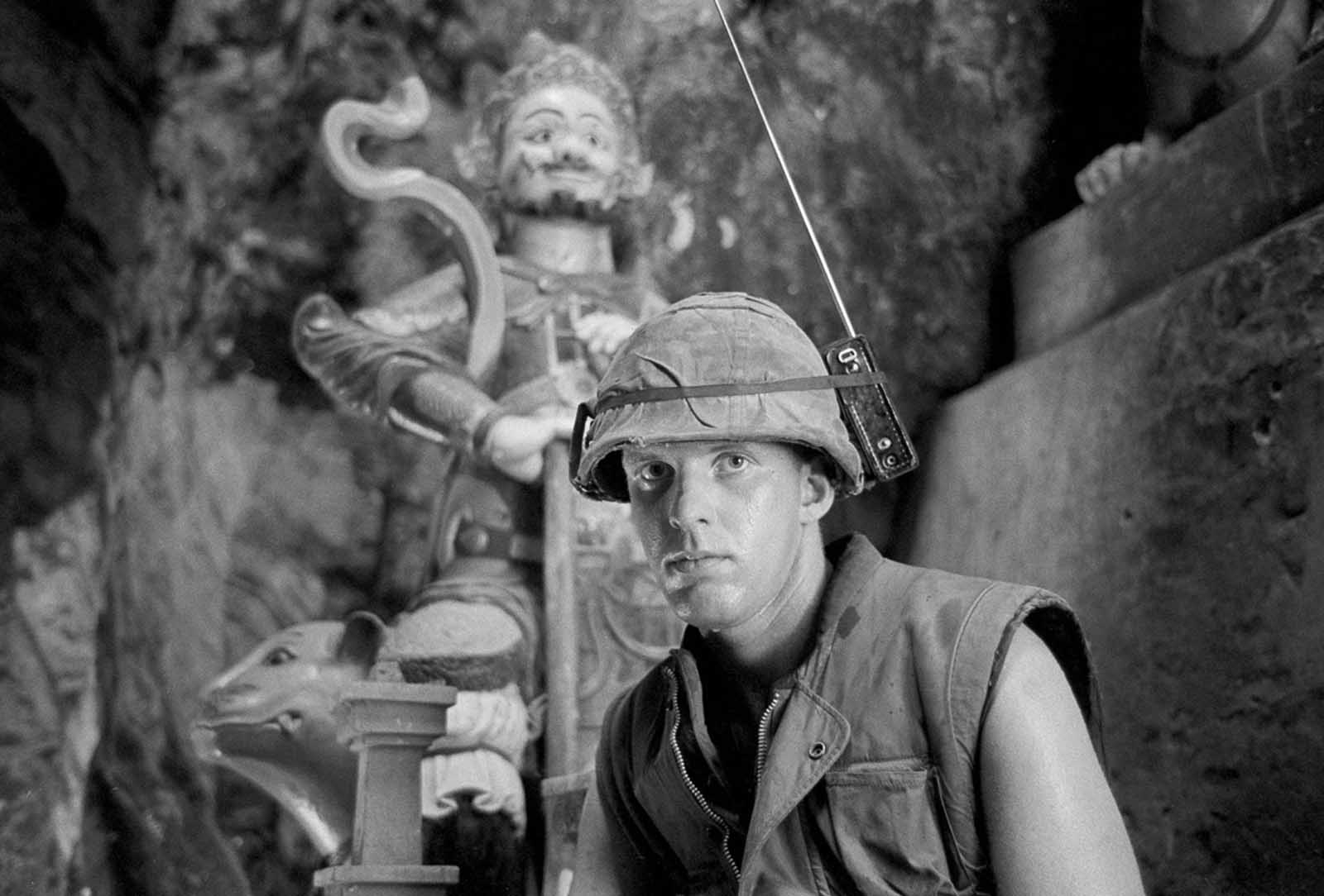 Marine Lance Corporal David L. Cruz tunes into the latest news on the Apollo moon shot on a helmet-mounted transistor radio while standing guard at Da Nang's Marble Mountain, on July 17, 1969. In background is a tall Buddhist figure found in many limestone caves of the mountain.