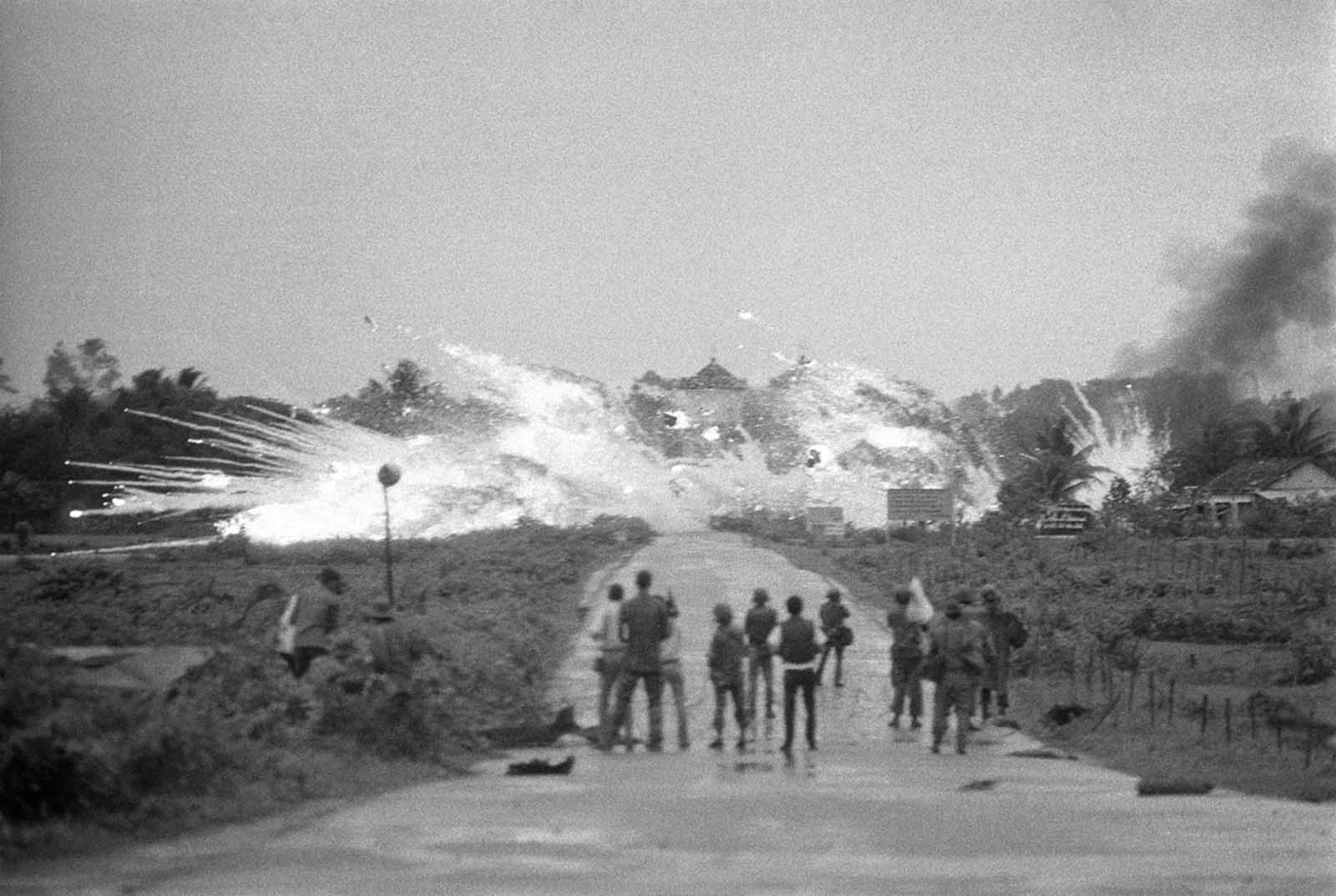 Bombs with a mixture of napalm and white phosphorus jelly dropped by Vietnamese AF Skyraider bombers explode across Route 1, amid homes and in front of the Cao Dai Temple in the outskirts of Trang Bang, on June 8, 1972. In the foreground are Vietnamese soldiers and news and cameramen from various international news organizations who watch the scene.
