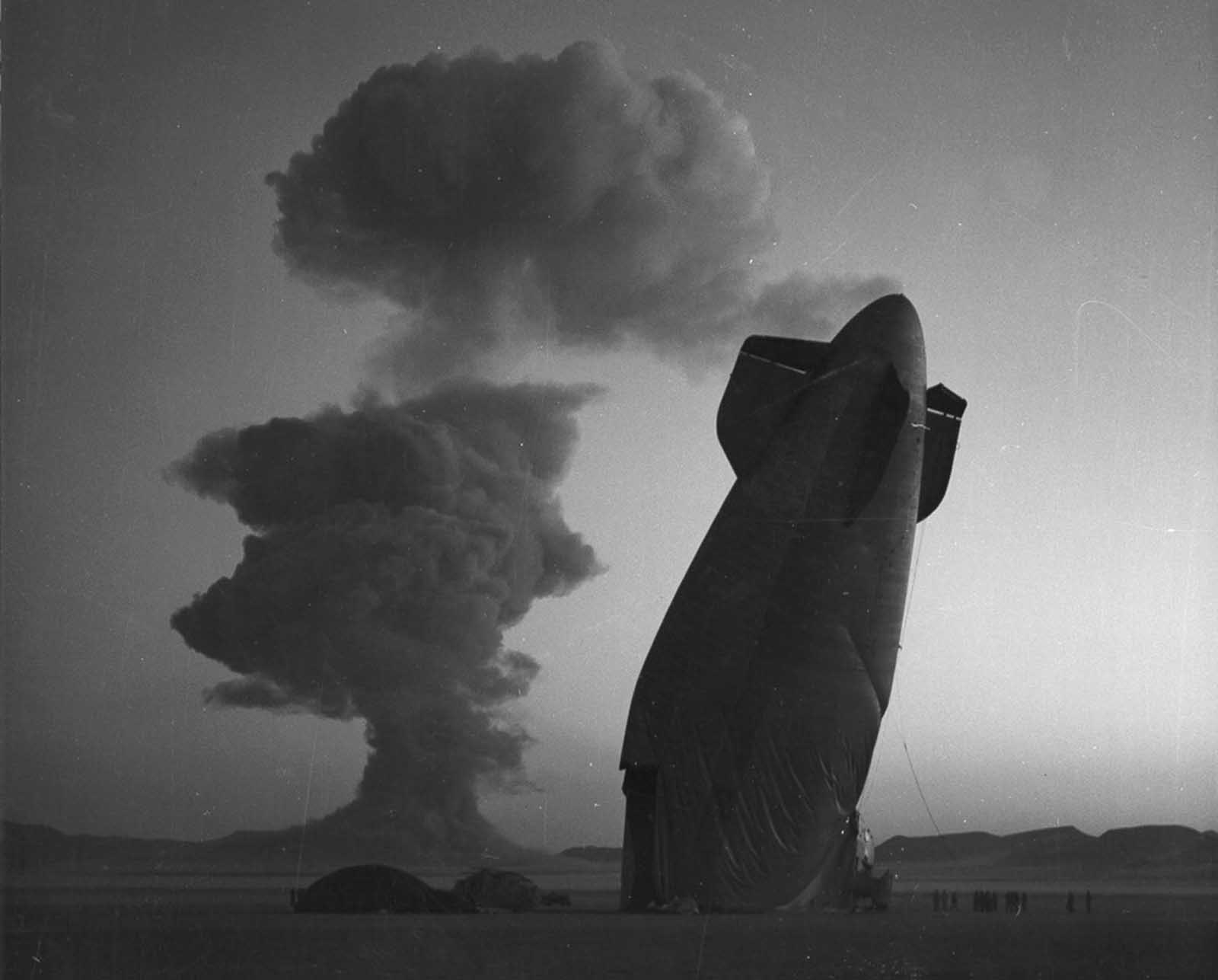 The tail section of a U.S. Navy blimp is shown with the Stokes cloud in the background at the Nevada Test Site on August 7, 1957. The blimp was in temporary free flight in excess of five miles from ground zero when it was collapsed by the shock wave from the blast. The airship was unmanned and was used in a military effects experiment.