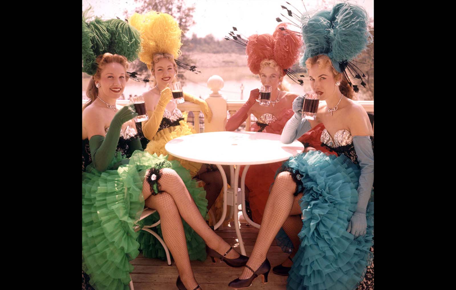 Showgirls in costume sit at an outdoor table and drink from mugs at Disneyland in July 1955.