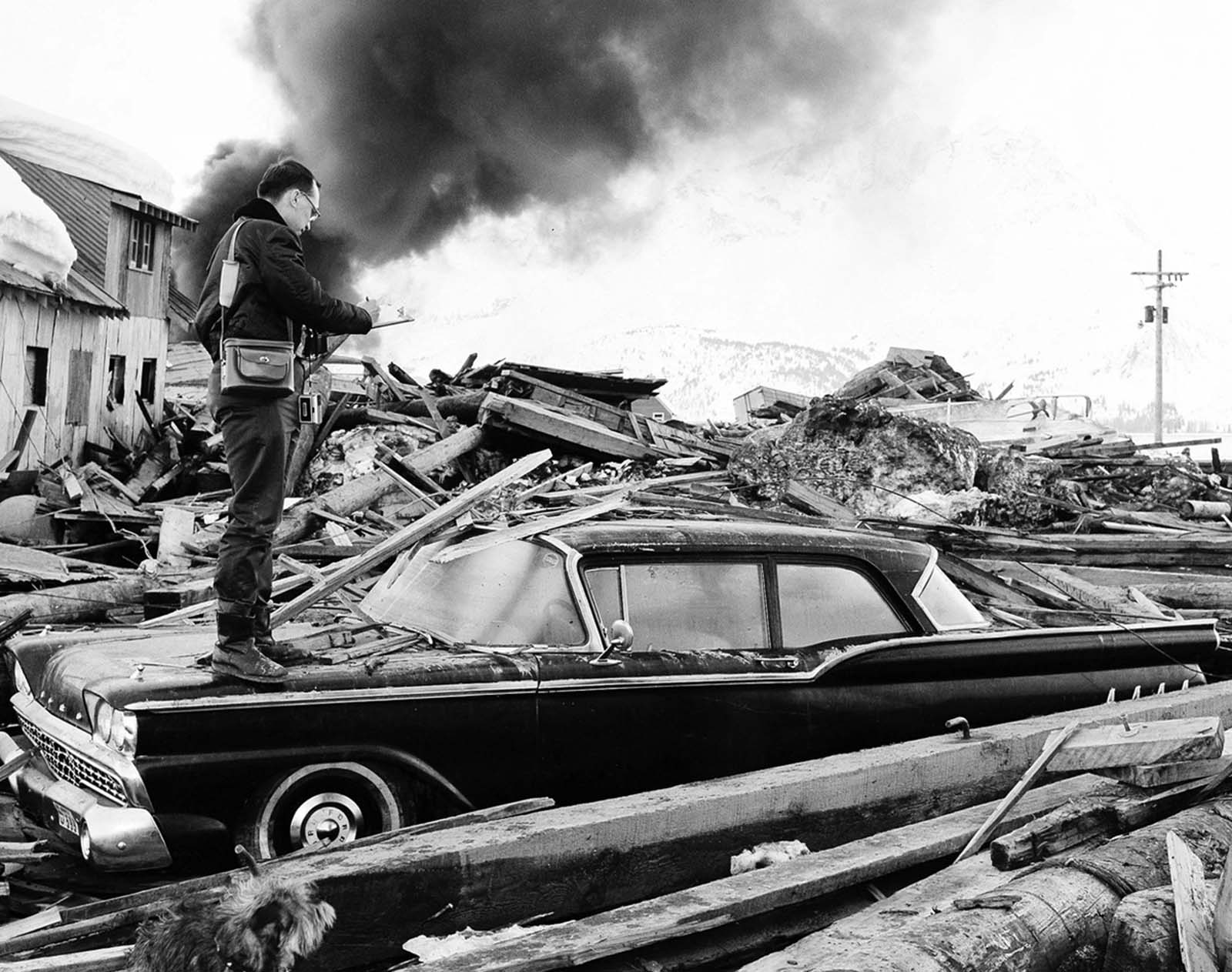 A photographer looks over wreckage as smoke rises in the background from burning oil storage tanks in Valdez, Alaska, on March 29, 1964, two days after the earthquake struck.