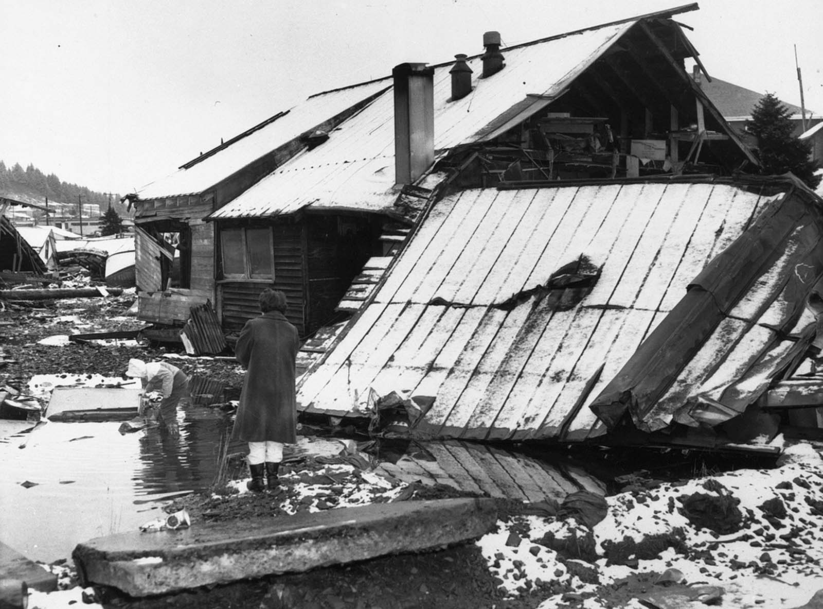 A mother stands watch as her child plays in a puddle of water near her earthquake-shattered home in Kodiak, Alaska in March of 1964.