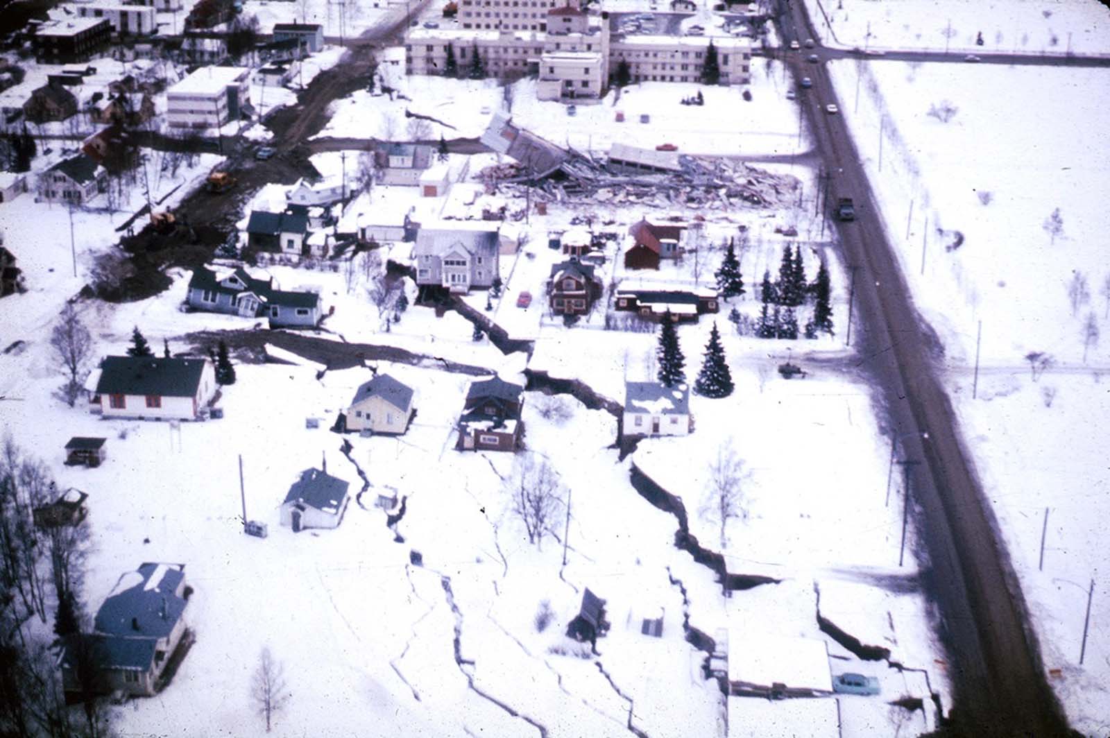 The head of the L Street landslide in Anchorage. The land on the left side sank 7 to 10 feet in response to 11 feet of horizontal movement of the lower section of the slide. A number of houses were undercut or tilted by subsidence of the graben. Note also the collapsed Four Seasons Apartment Building and the undamaged three-story reinforced concrete frame building behind it, which are on more stable ground.