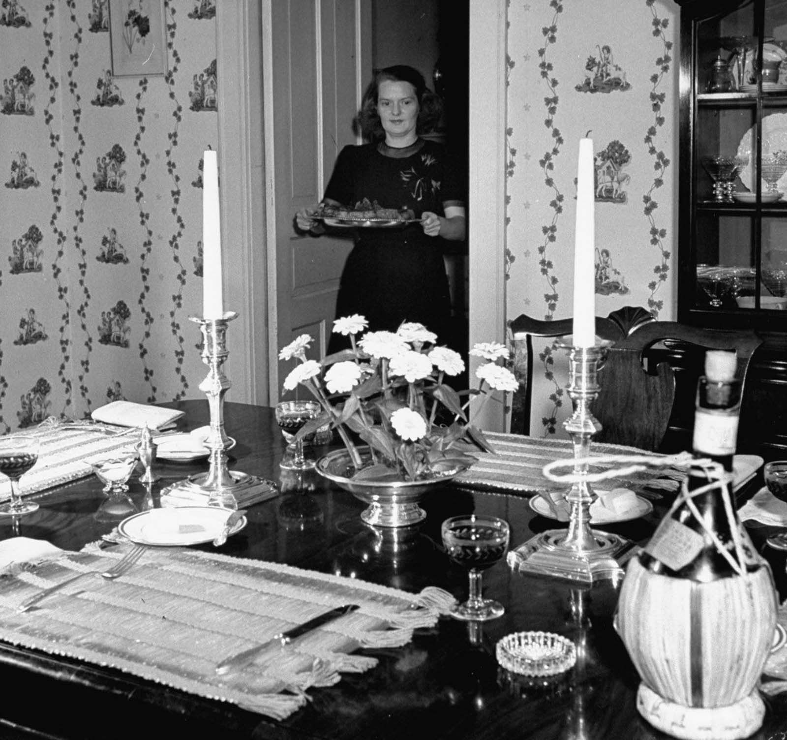 Jane prepares for dinner party by carrying a roast beef platter to the dining table she has carefully set with colorful placemats, silver candlesticks and silver centerpiece with flowers and a bottle of Chianti, as her guests finish cocktails in the living room.