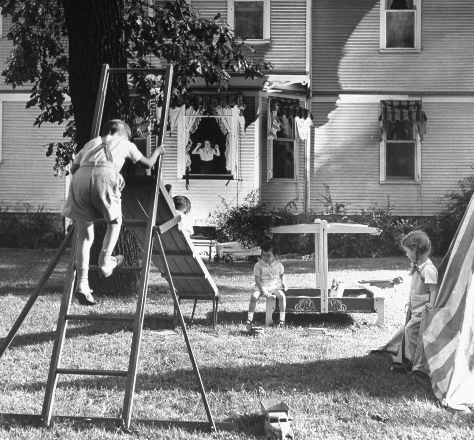 Jane rapping admonishingly on the window in the background as she oversees her kids Peter, 7, climbing the slide ladder, while Tony, 5, blocks the slide, and Pamela gets kicked out of the tent while playing with a neighbor's children.