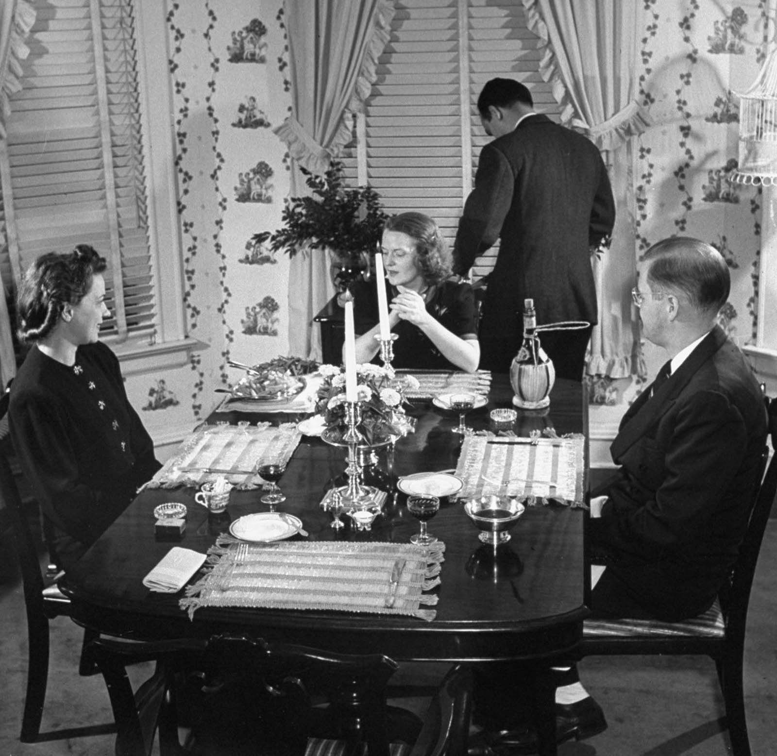 Jane chatting with her dinner guests Mr. Bert Miller and wife, as her husband Gilbert stands behind her, carving a roast beef to be served at the table.