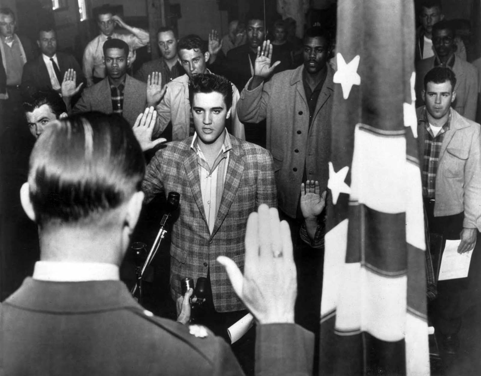 Elvis Presley stands with a group of young men at an induction center, raising their right hands as they are sworn into the United States Army by an officer standing next to an American flag.