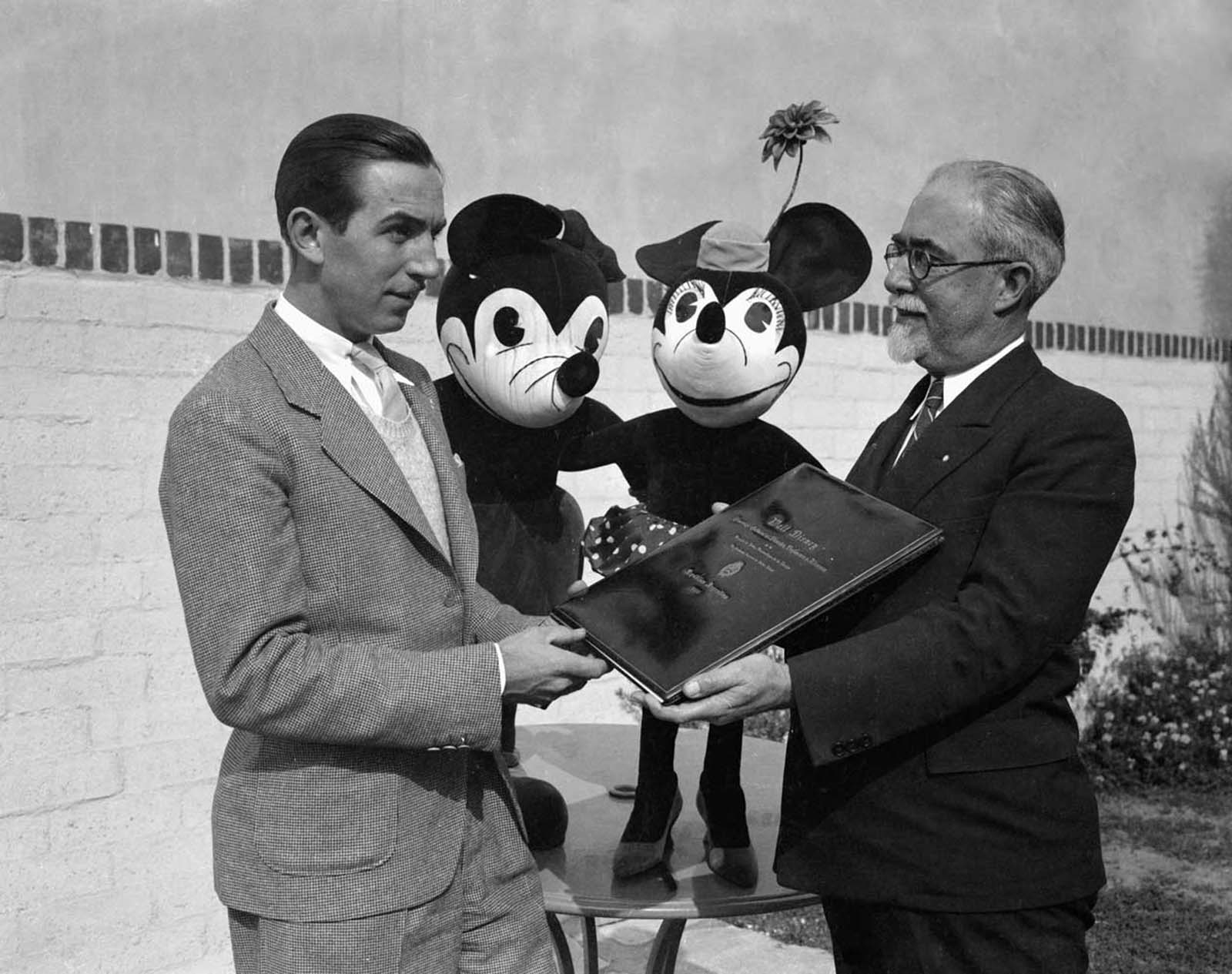 Walt Disney presented with a diploma of commendation from the National Academy of Fine Arts of Buenos Aires, Argentina. Mickey and Minnie look on.