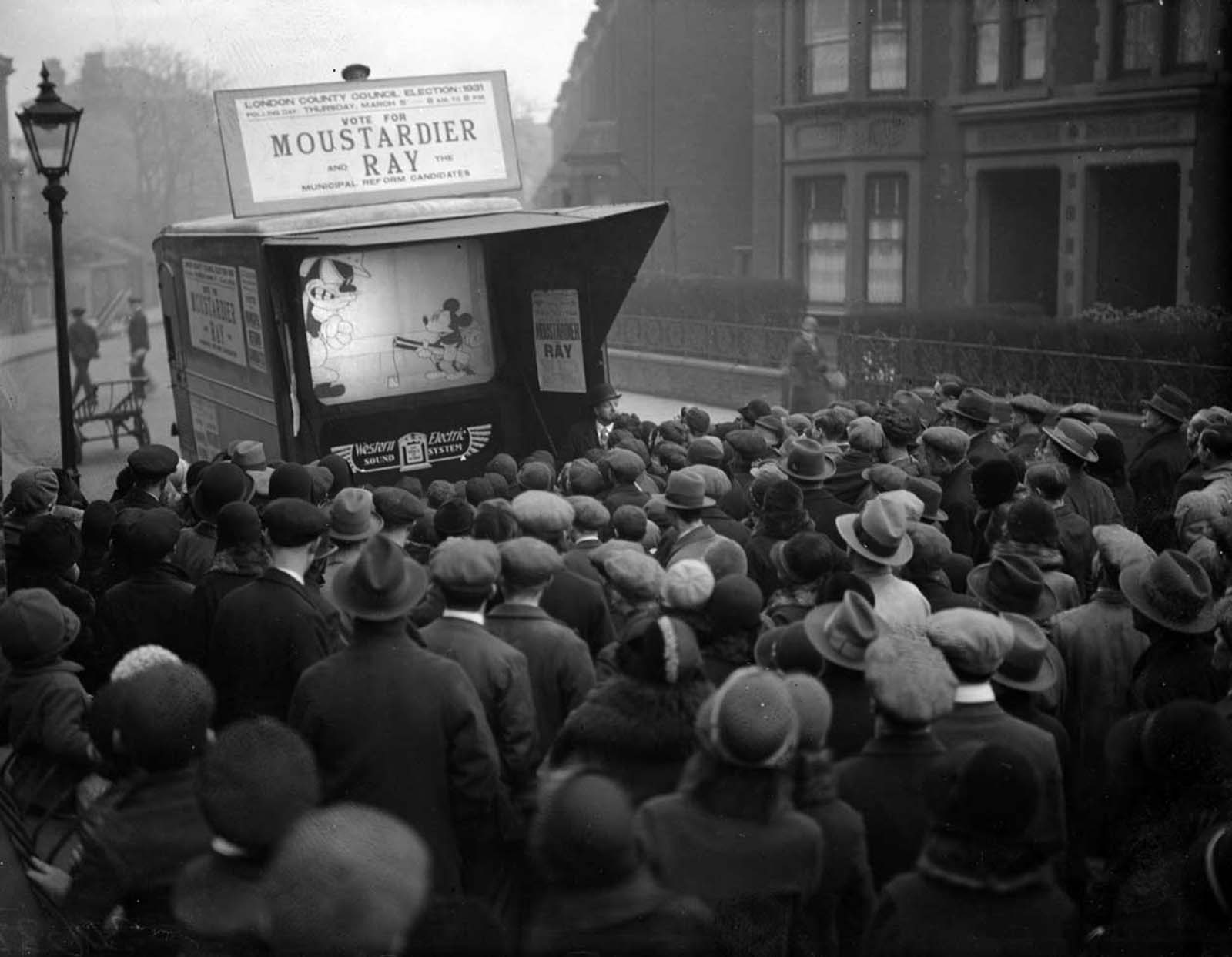 During a London County Council election campaign run, Mickey Mouse is showing on a small screen in a city street. 1931.