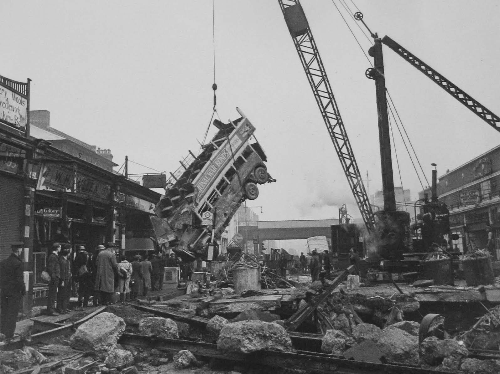 People are still moving along the footway beneath as the double decker bus is removed from the bomb crater.