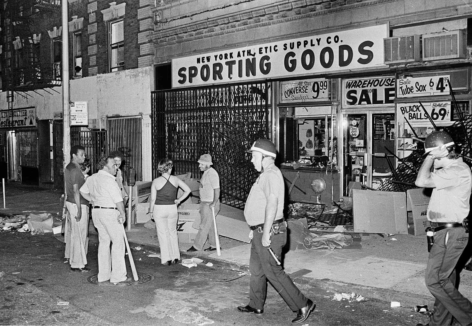 Owners and employees of a sporting goods store stand guard outside with baseball bats after the store on New York City's Upper West Side was looted during massive power failure, Wednesday night, July 13, 1977. Police patrolling the area walk by the store.