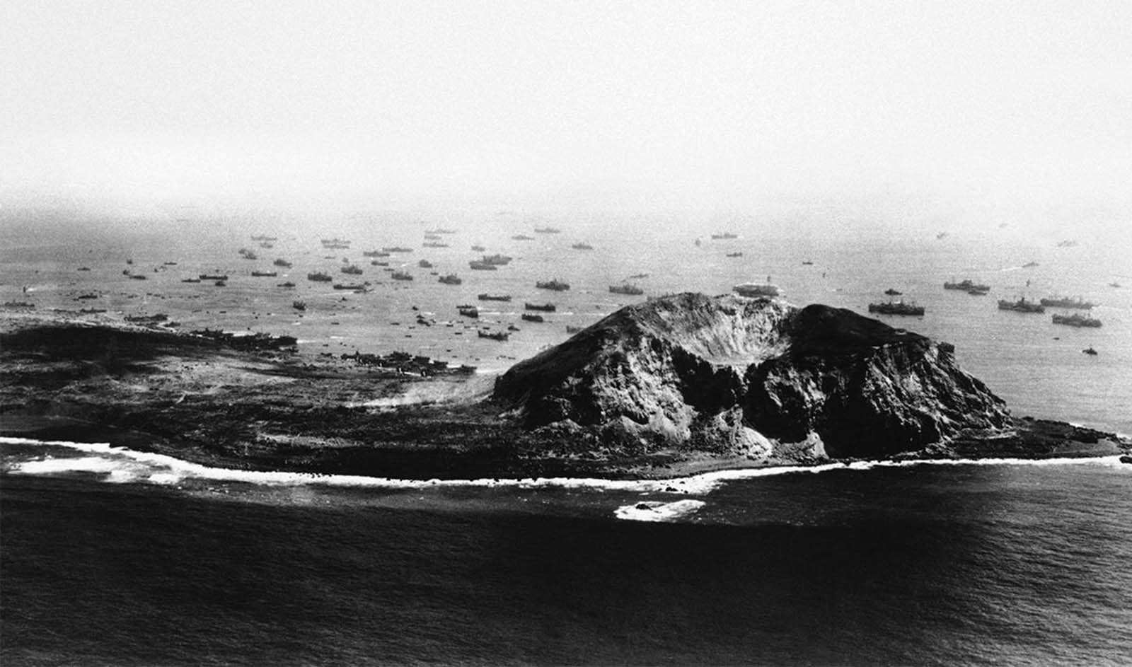 This aerial photograph made on day five of the invasion shows the immense power needed to break the back of Japanese Resistance on Iwo Jima, on March 17, 1945. Just off the beach, landing craft await their chance at the unloading area while small boats from the transports ply back and forth bring assault troops and returning wounded for treatment. Further out, the transports themselves faintly along the horizon, the protective screen of destroyers, destroyer escorts and cruisers can be seen. On the island, Marine tanks can be spotted moving through the rough terrain toward the first airfield at left.
