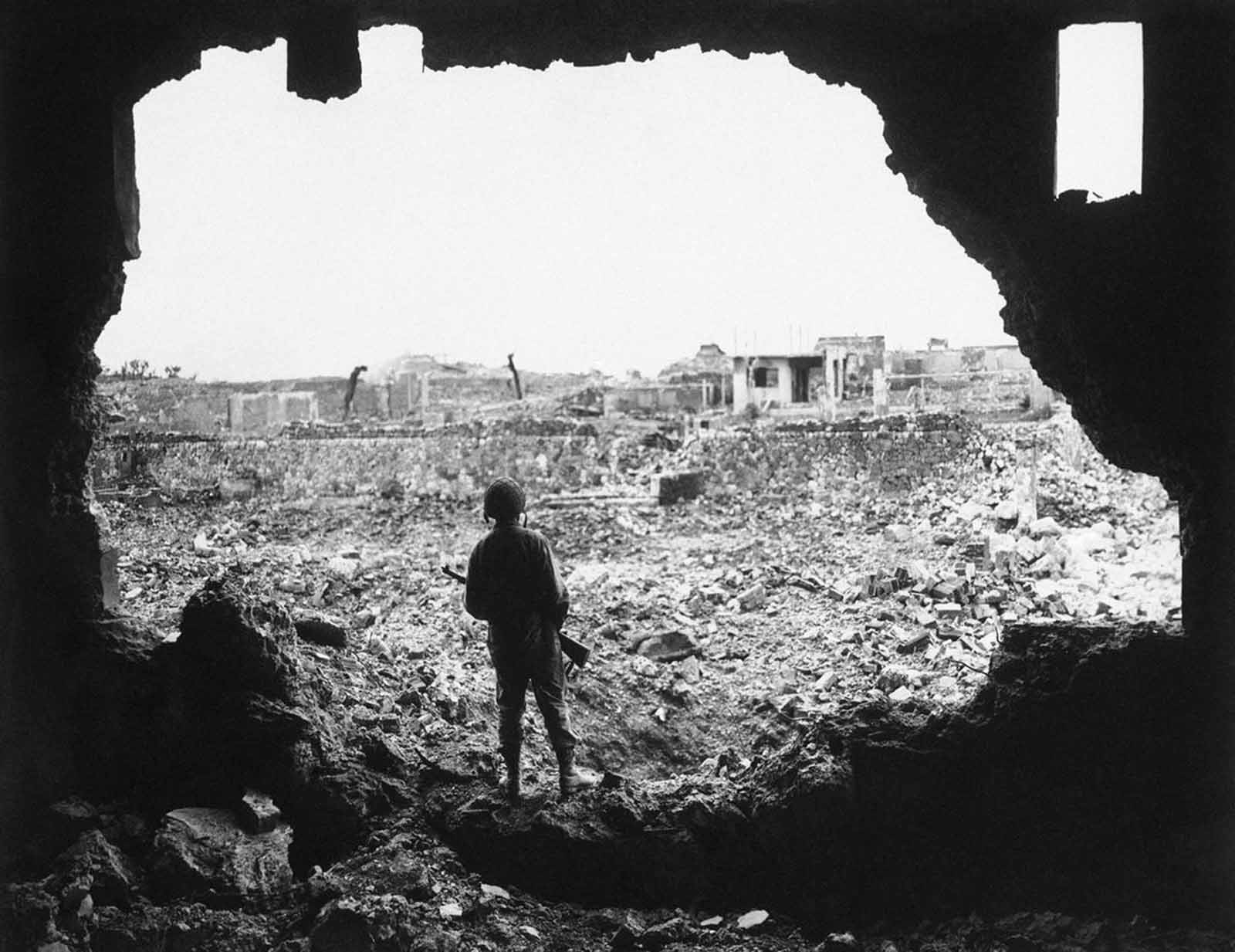 Perched on the rim of a gaping hole in the wall of a theater in the Ryukyu capital, a Marine rifleman views the result of the American bombardment of Naha, Okinawa, Japan, on June 13, 1945. Structure skeletons are all that remain of the city with a pre-invasion population of 443,000 people.