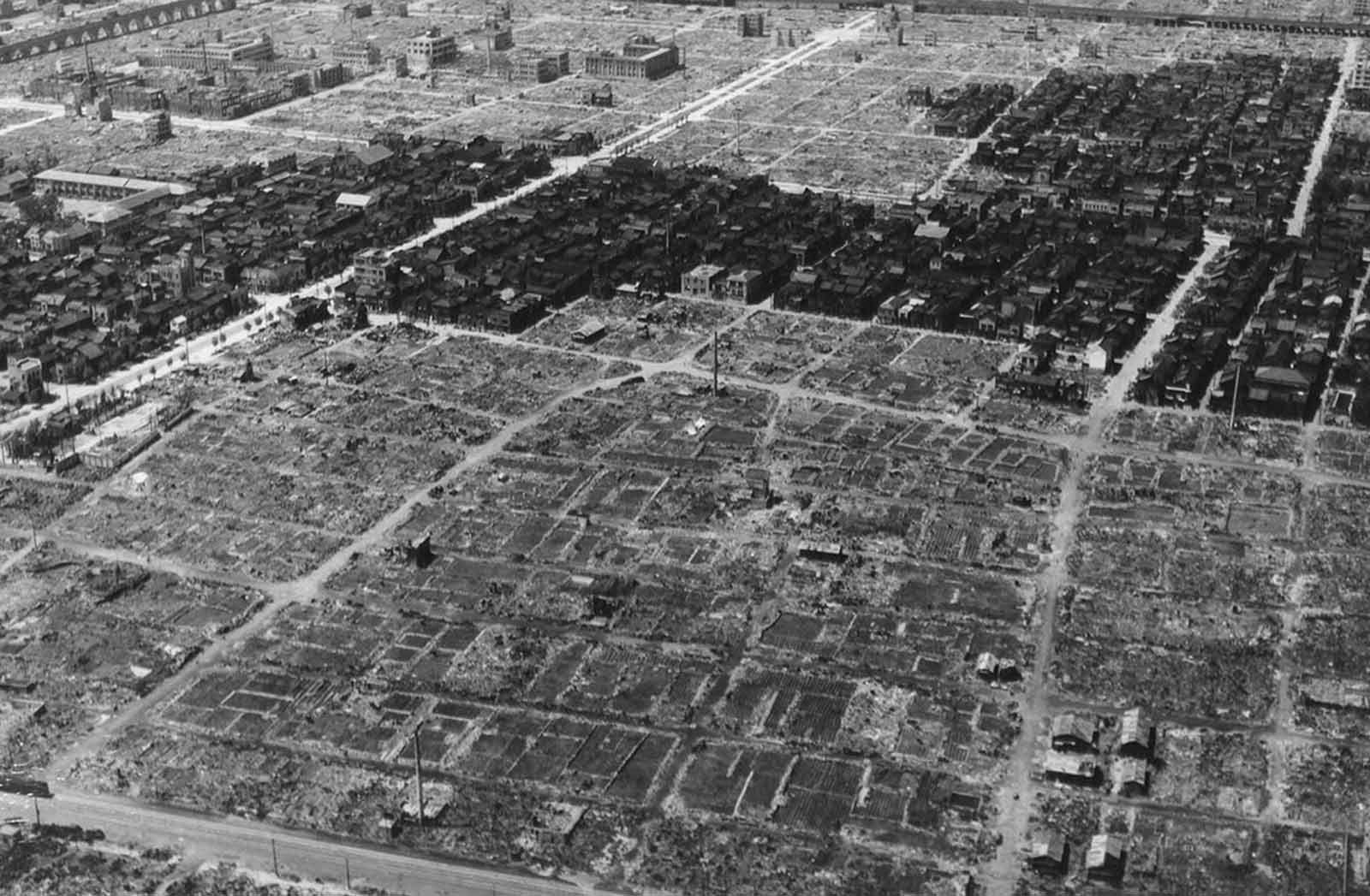After an incendiary bombing, a view of some of the damage in Tokyo, Japan in 1945. A strip of residential buildings remains undamaged, surrounded by ashes and rubble of neighboring structures burned or blasted to the ground.