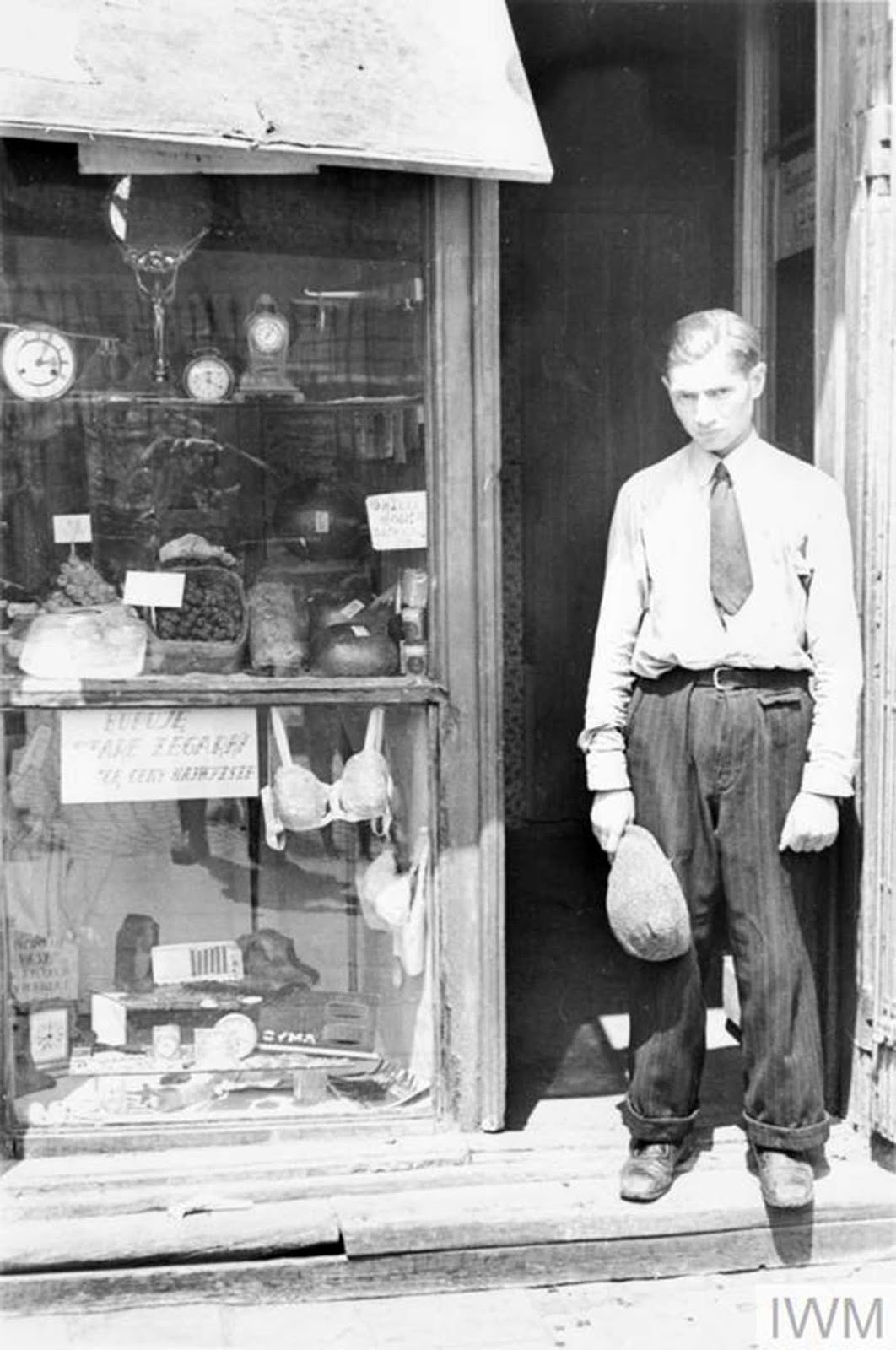 A young man in the doorway of a shop in the ghetto. Note he has taken his hat off to comply with the German order to remove headwear in the presence of German personnel. The shop offers fresh eggs, sweets and watches. The sign in the window reads - 
