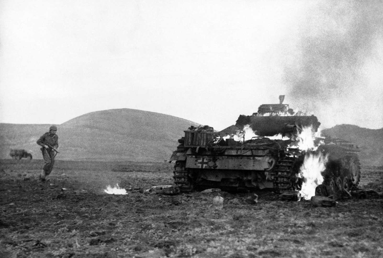 A United States soldier advances cautiously at left with a sub-machine gun to cover any attempt of the German tank crew from escaping their fiery prison inside their tank following a duel with U.S. and British anti-tank units in Medjez al Bab area, Tunisia, on January 12, 1943.