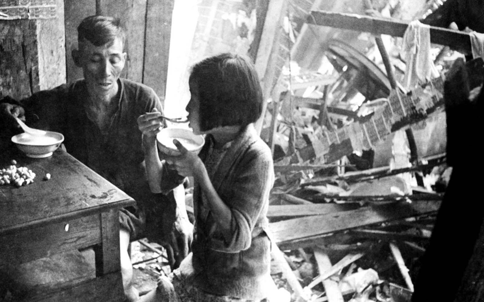 Dining together after a Japanese raid in the Singapore area, on February 26, 1942. A Chinese man and his young daughter sit calmly amid the ruins eating bowls of rice.