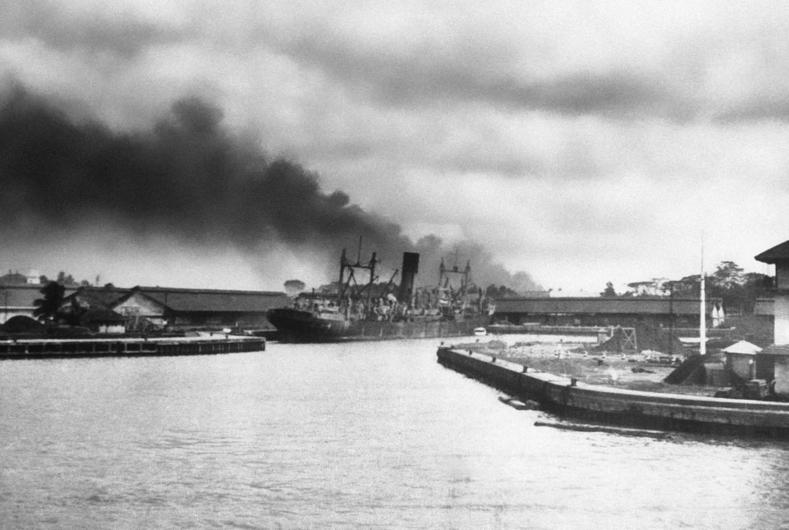 A large freighter settles slowly after being hit by Japanese bombs alongside of one of Singapore's docks on February 12, 1942. Smoke from other struck objectives billows over the waterfront in this photo by C. Yates McDaniel, Associated Press correspondent, who was among the last to leave the besieged port on February 12. The next day his ship was bombed and he reached safety after further harrowing experience.