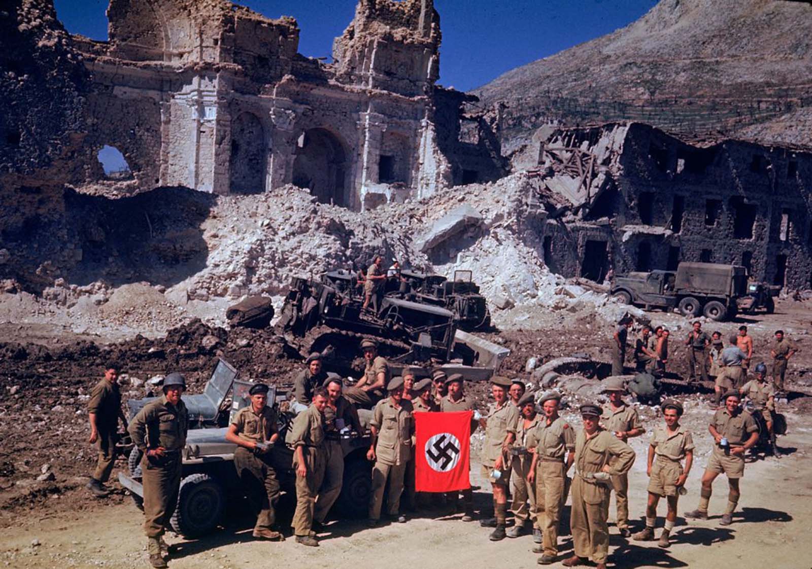 British and South African soldiers held up a Nazi trophy flag while combat engineers on bulldozers cleared a path through the debris of a bombed-out city, Italian Campaign, World War II.