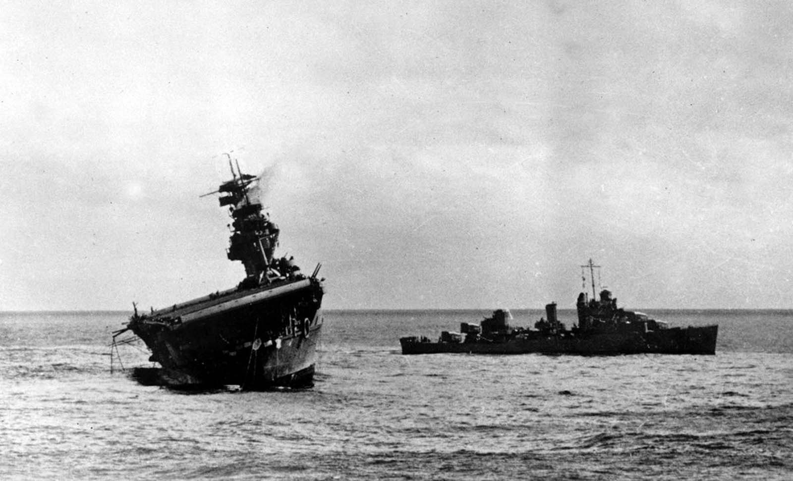 The USS Yorktown lists heavily to port after being struck by Japanese bombers and torpedo planes in the Battle of Midway on June 4, 1942. A destroyer stands by at right to assist as a salvage crew on the flight deck tries to right the stricken aircraft carrier.