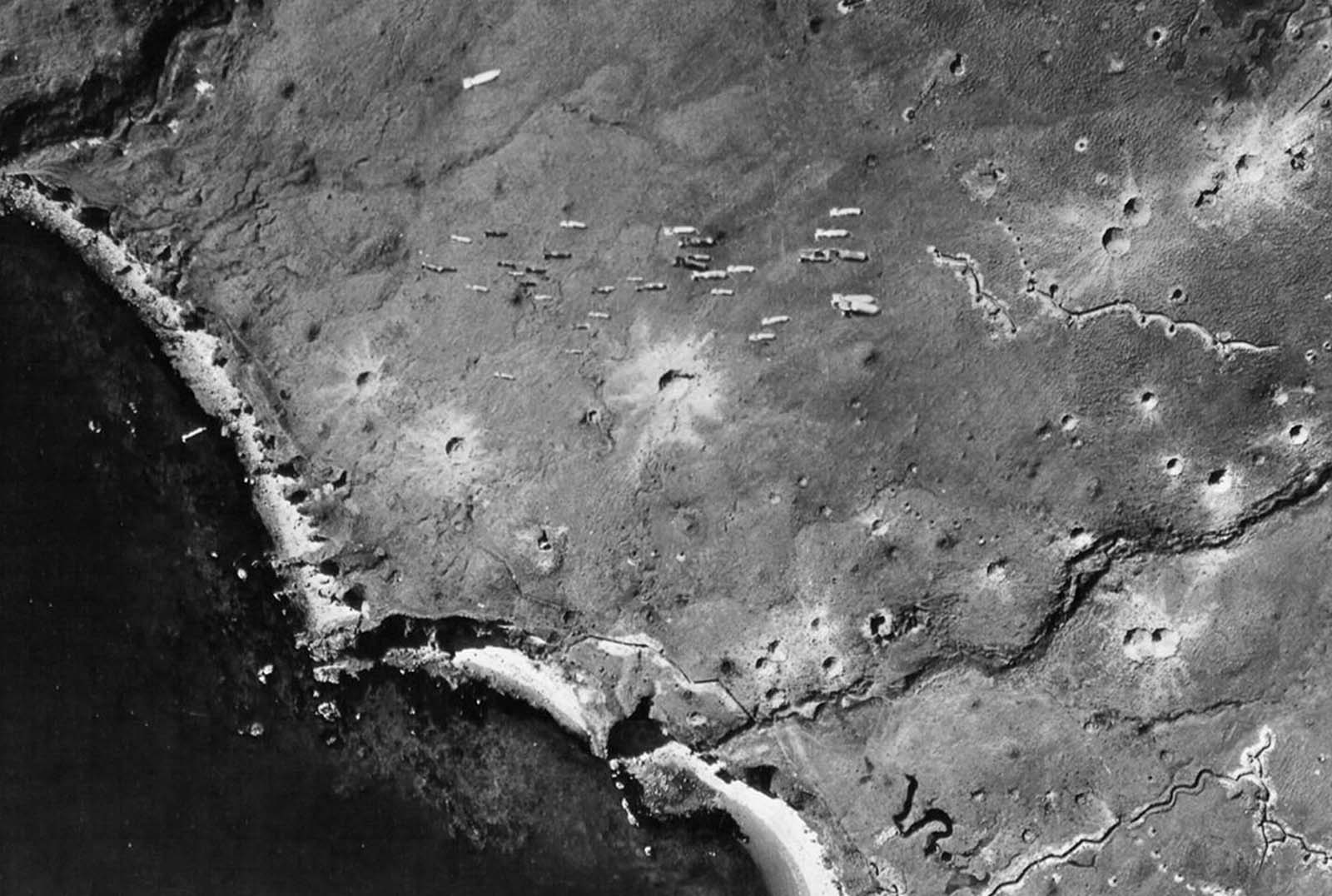 Dozens of bombs fall from a U.S. bomber toward Japanese-occupied Kiska Island, Alaska, on August 10, 1943. Note the craters from previous bombing runs and the zig-zag trenches dug by the Japanese.
