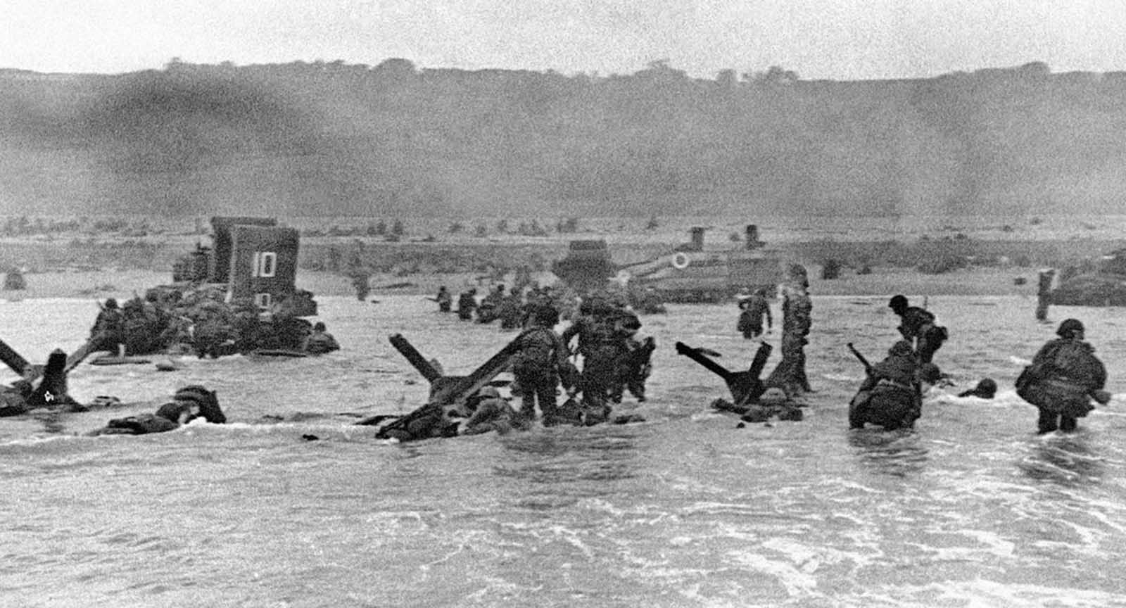 Some of the first assault troops to hit the beachhead in Normandy, France take cover behind enemy obstacles to fire on German forces as others follow the first tanks plunging through the water towards the German-held shore on June 6, 1944.