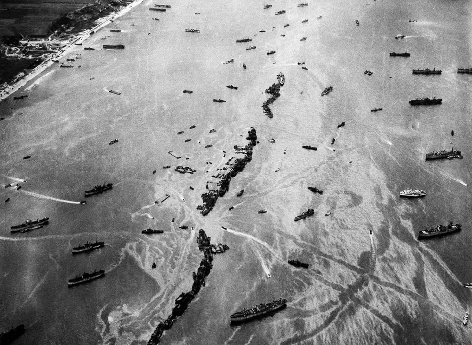 Thirteen liberty ships, deliberately scuttled to form a breakwater for invasion vessels landing on the Normandy beachhead lie in line off the beach, shielding the ships in shore. The artificial harbor installation was prefabricated and towed across the Channel in 1944.