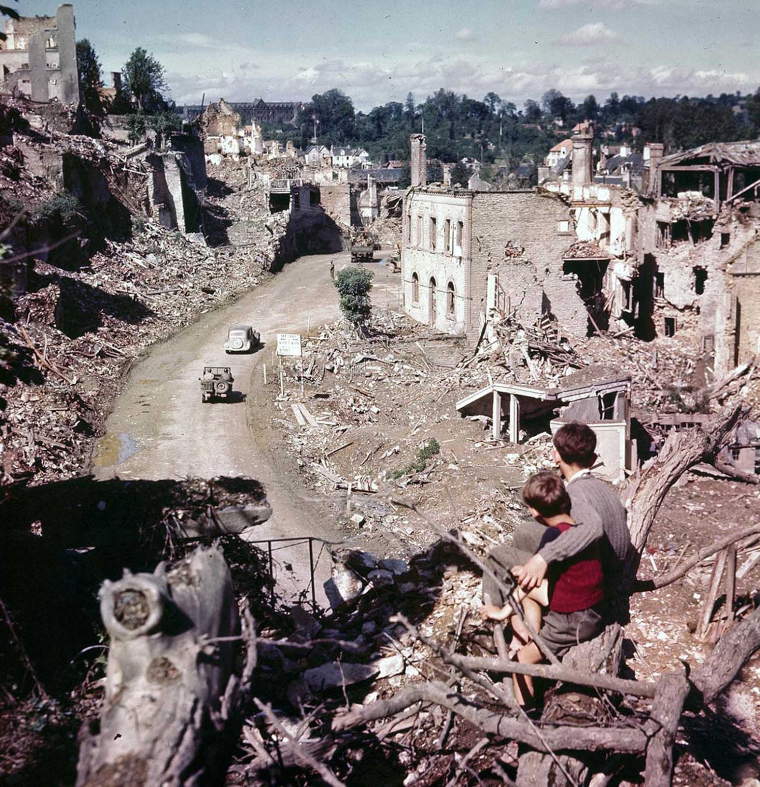 A view from a hilltop overlooking the road leading into St. Lo in July of 1944. Two French children in the foreground watch convoys and trucks of equipment go through their almost completely destroyed city en route to the front.