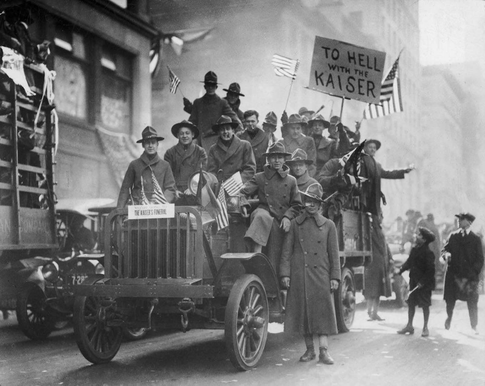 A group of American soldiers ride in a truck, waving American flags during an Armistice Day parade , New York City. One soldier holds a sign reading 'To Hell With The Kaiser.'