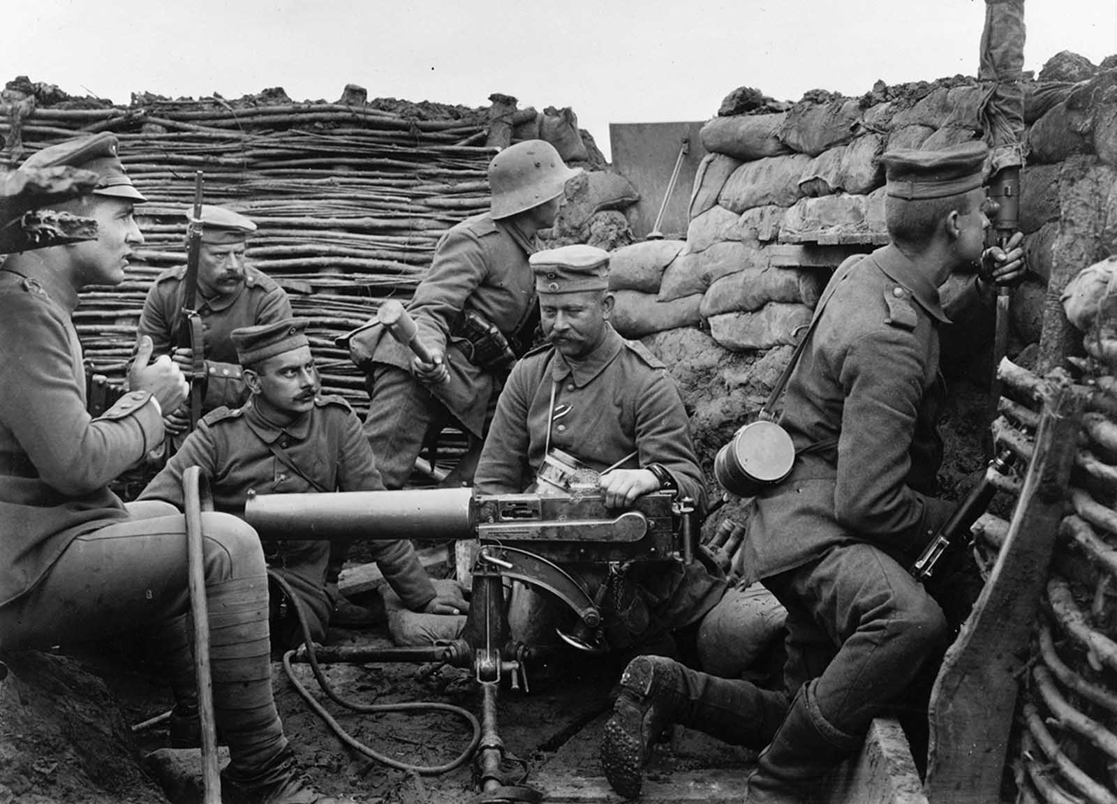 Six German soldiers pose in a in trench with machine gun, a mere 40 meters from the British line, according to the caption provided. The machine gun appears to be a Maschinengewehr 08, or MG 08, capable of firing 450-500 rounds a minute. The large cylinder is a jacket around the barrel, filled with water to cool the metal during rapid fire. The soldier at right, with gas mask canister slung over his shoulder, is peering into a periscope to get a view of enemy activity. The soldier at rear, with steel helmet, holds a