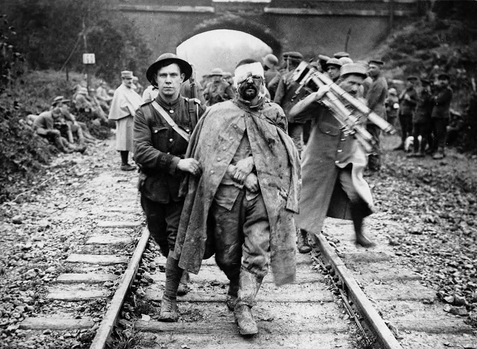 A German prisoner, wounded and muddy, helped by a British soldier along a railway track. A man, possibly in French military uniform, is shown behind them, holding a camera and tripod, ca. 1916.