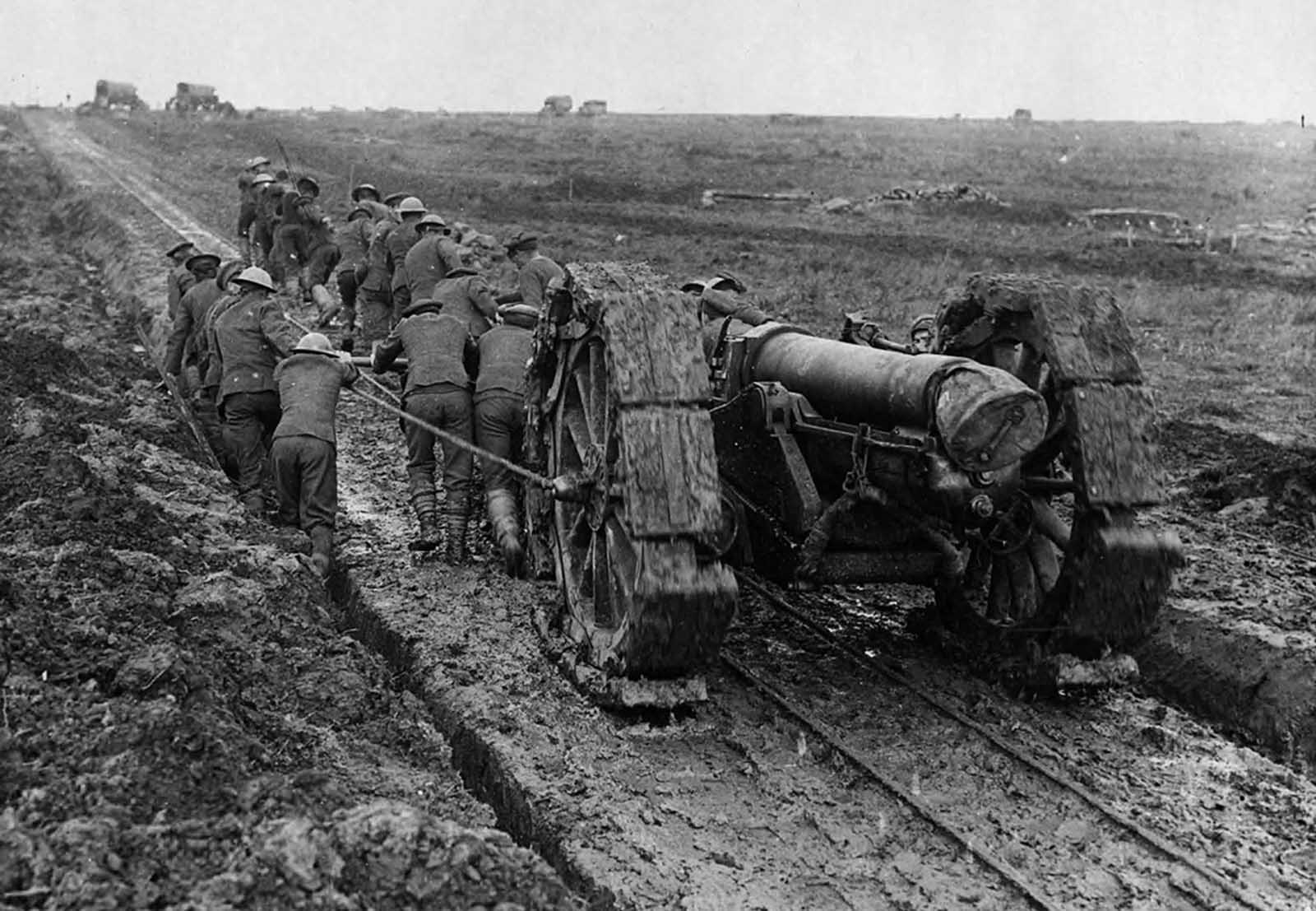 Soldiers struggle to pull a huge piece of artillery through mud. The gun has been placed on a track created for a light railway. The soldiers are pushing a device, attached to the gun, that possibly slots into the tracks. Some of the men are in a ditch that runs alongside the track, the rest are on the track itself. A makeshift caterpillar tread has been fitted to the wheels of the gun, in an attempt to aid its movement through the mud.