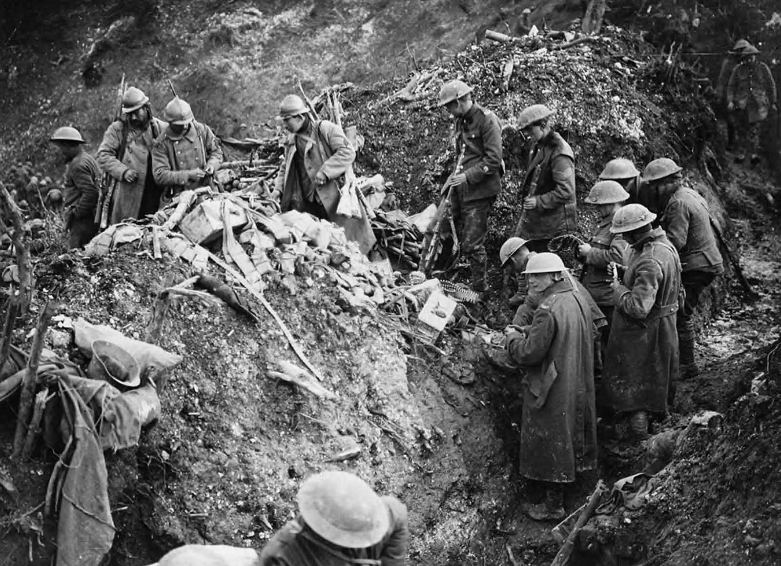 Cleaning up German trenches at St. Pierre Divion. In the foreground a group of British soldiers are sorting through equipment abandoned in the trenches by the Germans when St Pierre Divion was captured. One soldier has three rifles slung on his shoulder, another has two. Others are looking at machine gun ammunition. The probable photographer, John Warwick Brooke, has achieved considerable depth of field as many other soldiers can be seen in the background far along the trenches.