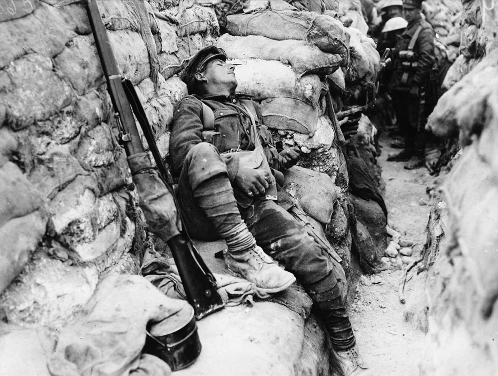 Soldier's comrades watch him as he sleeps, near Thievpal, France. Soldiers are standing in a very deep, narrow trench, the walls of which are entirely lined with sandbags. At the far end of the trench a line of soldiers are squashed up looking over each others' shoulders at the sleeping man.